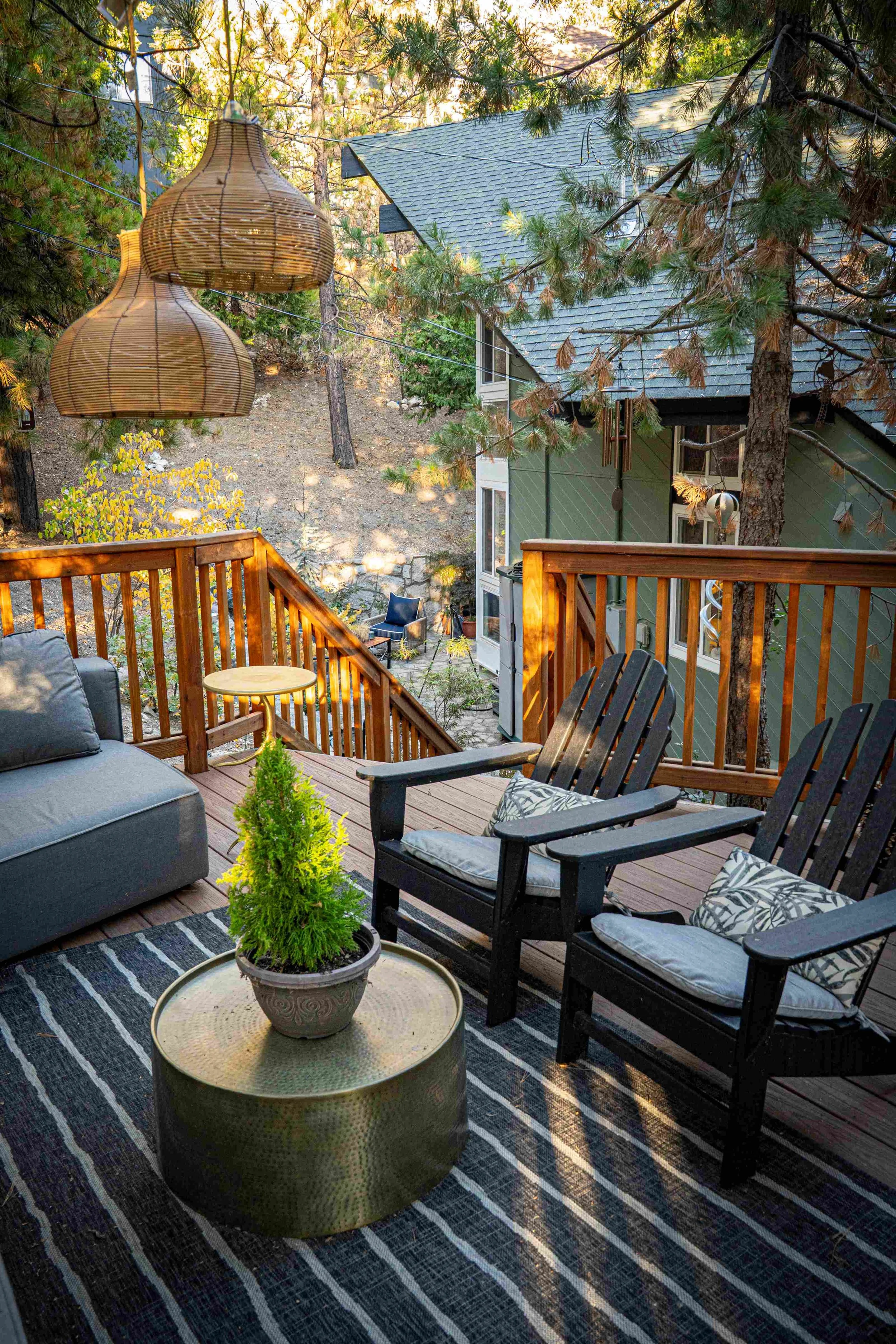A cozy outdoor deck with a black and white striped rug, two black wooden armchairs with patterned cushions, a gray sofa, a small round table with a potted plant, hung bamboo pendant lights, surrounded by trees and overlooking a backyard with outdoor 