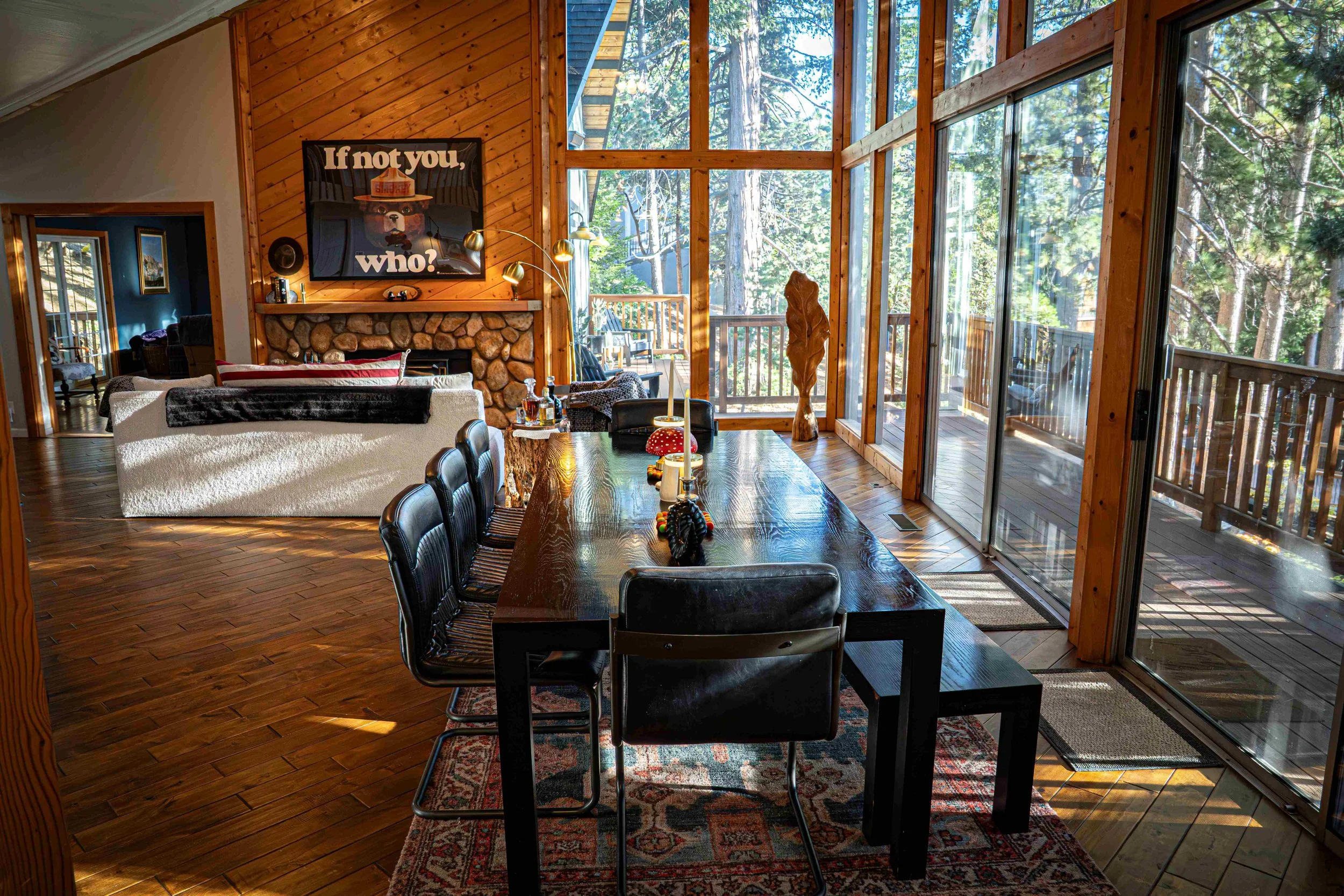 Interior of a cozy wooden cabin with a glass sunroom, a dining table with chairs, a living room with a sofa, and a fireplace with a bear-themed art piece above it, surrounded by trees visible through the large windows.
