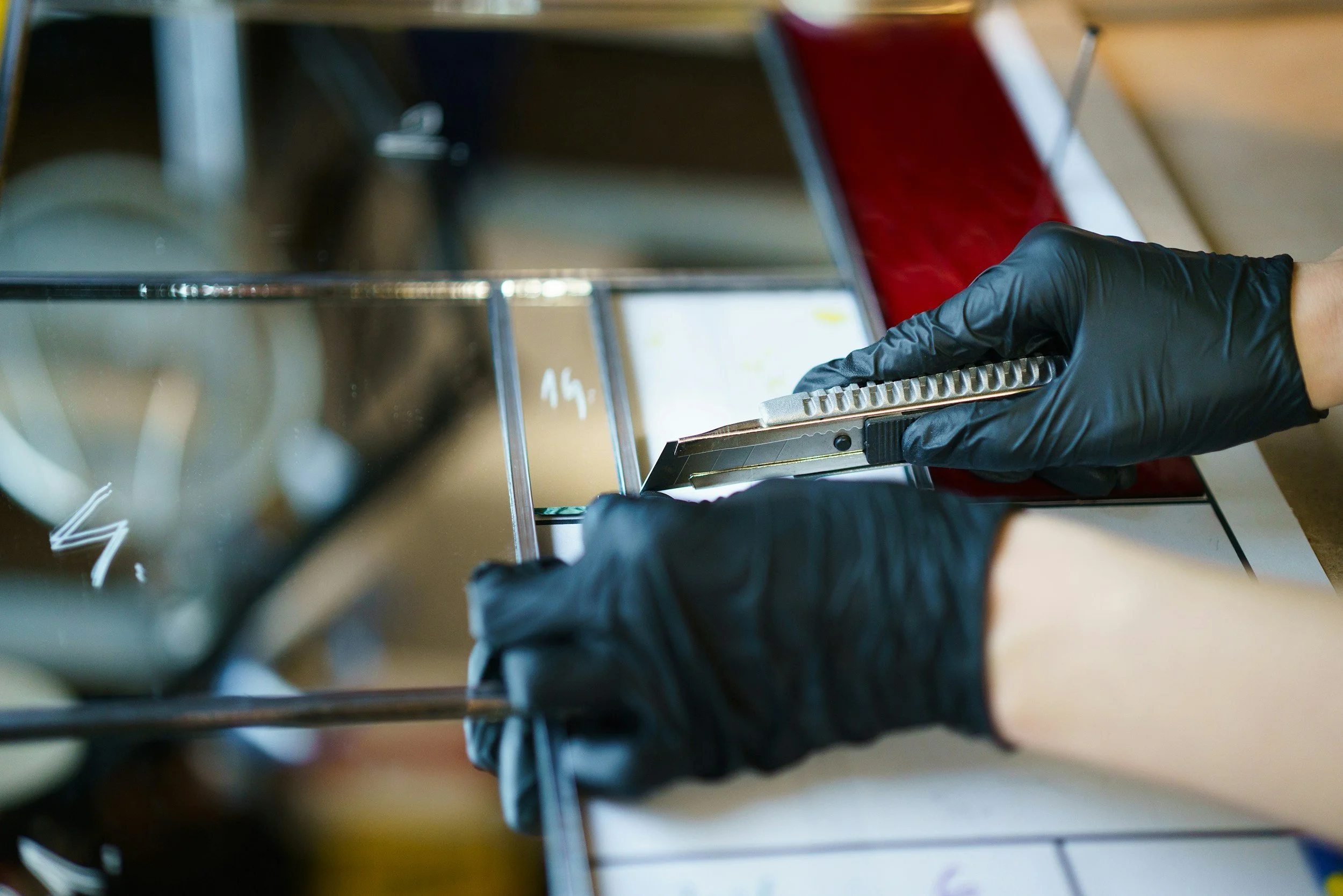 Person wearing black gloves using a metal tool to cut glass in a workshop.