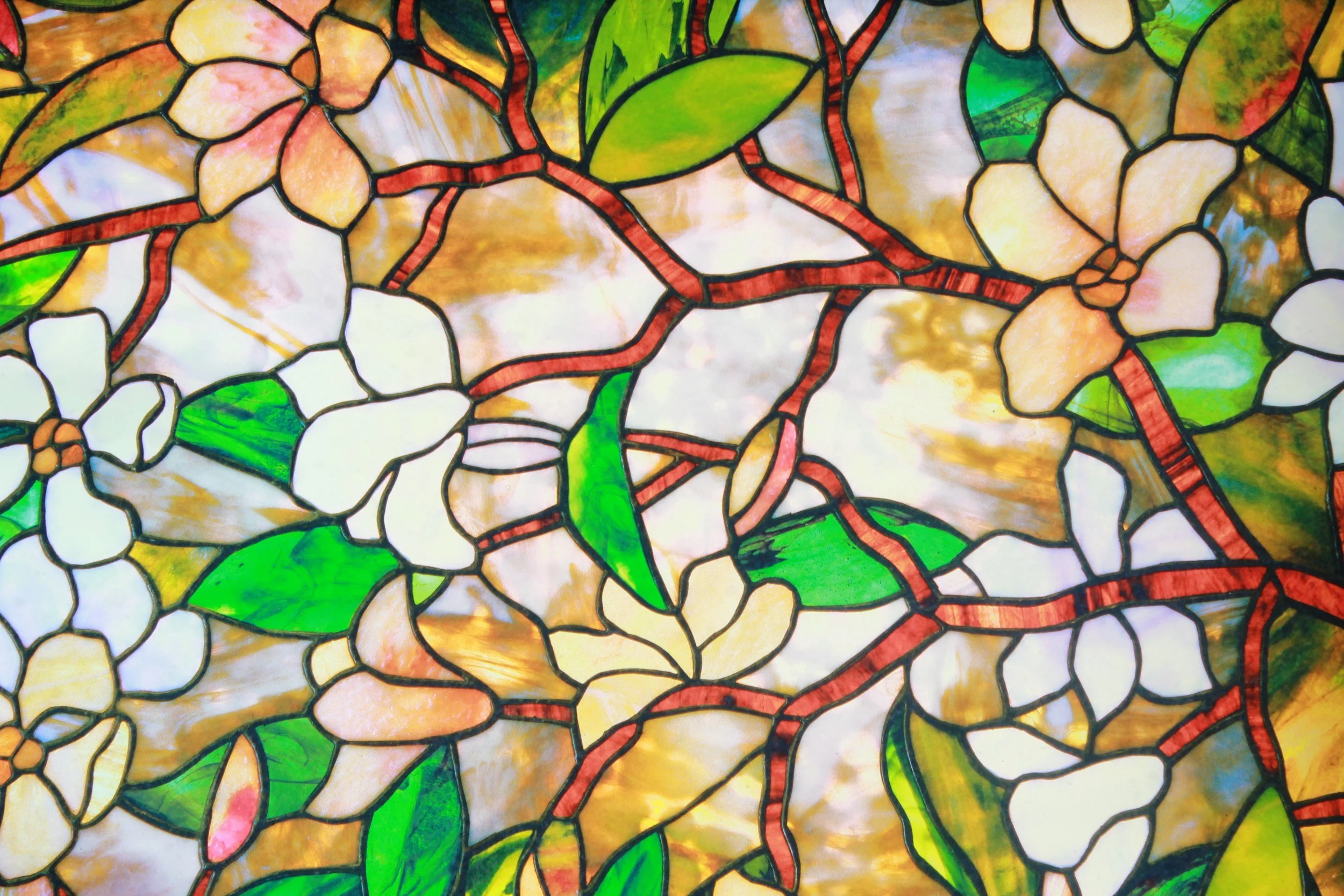 Close-up of stained glass window featuring white flowers, green leaves, and brownish-red branches with a multicolored background.