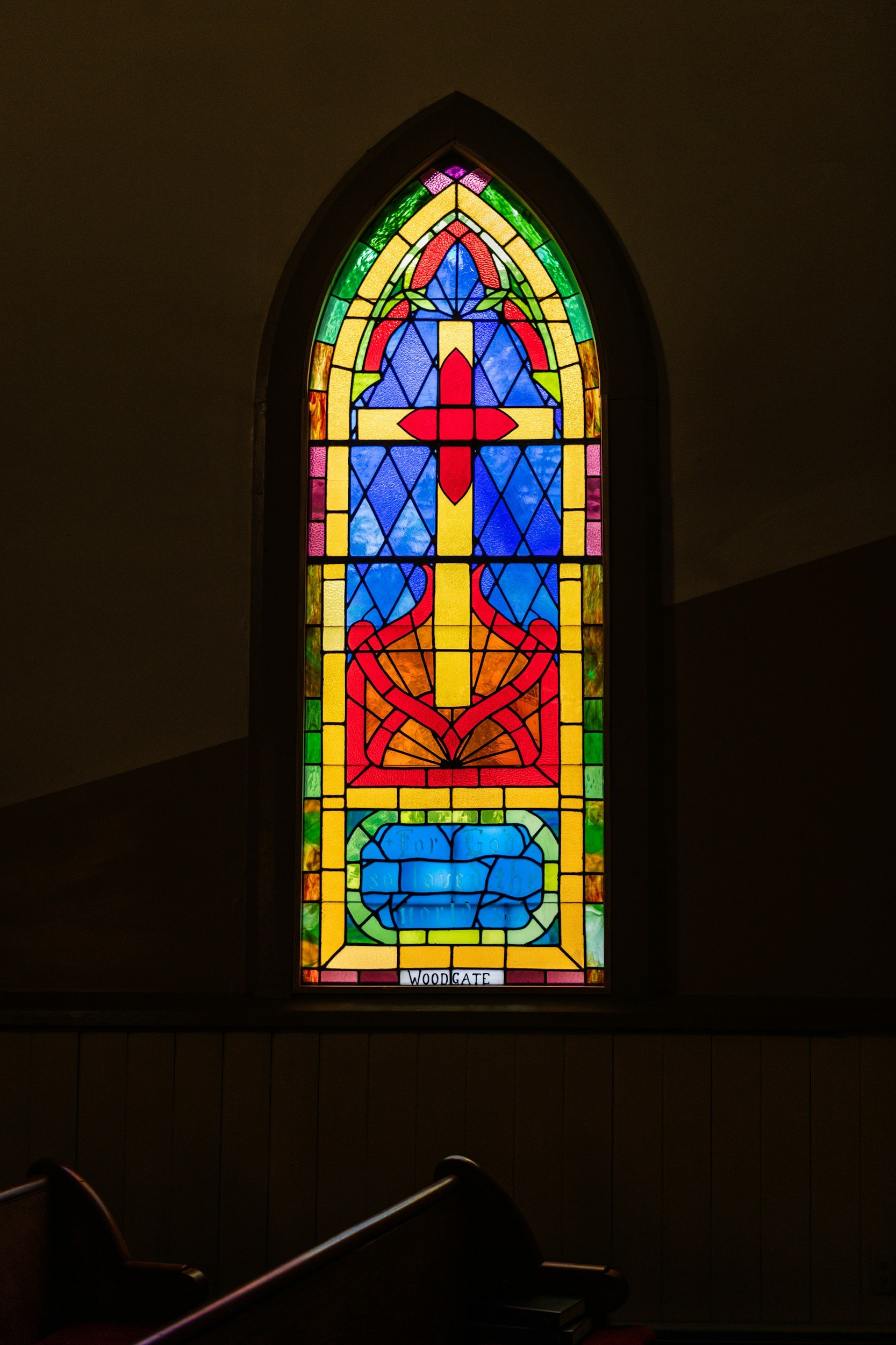 Colorful stained glass church window with a red cross, a sword, and a blue banner with the words 'For God and Country' at the bottom, labeled 'Woodgate'.