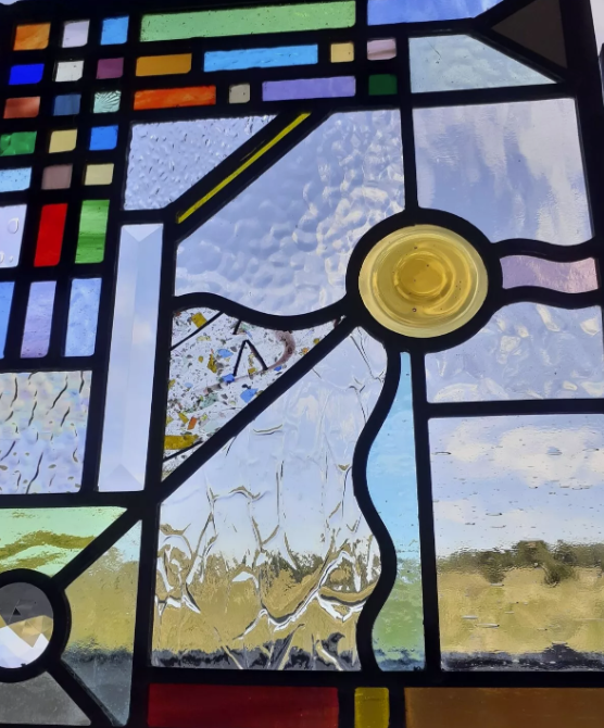 Close-up view of stained glass window featuring geometric shapes and a circular yellow glass element, with a scenic outdoor landscape visible through the glass.