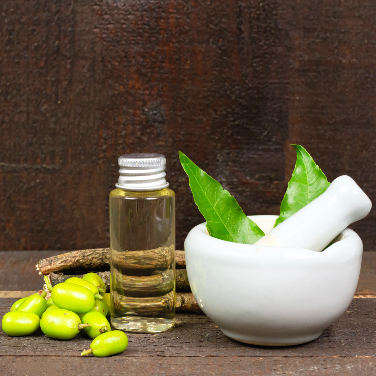 A small glass bottle of herbal oil, green jujube fruits, a white mortar and pestle with green leaves, all placed on a rustic wooden surface with a dark wooden background.