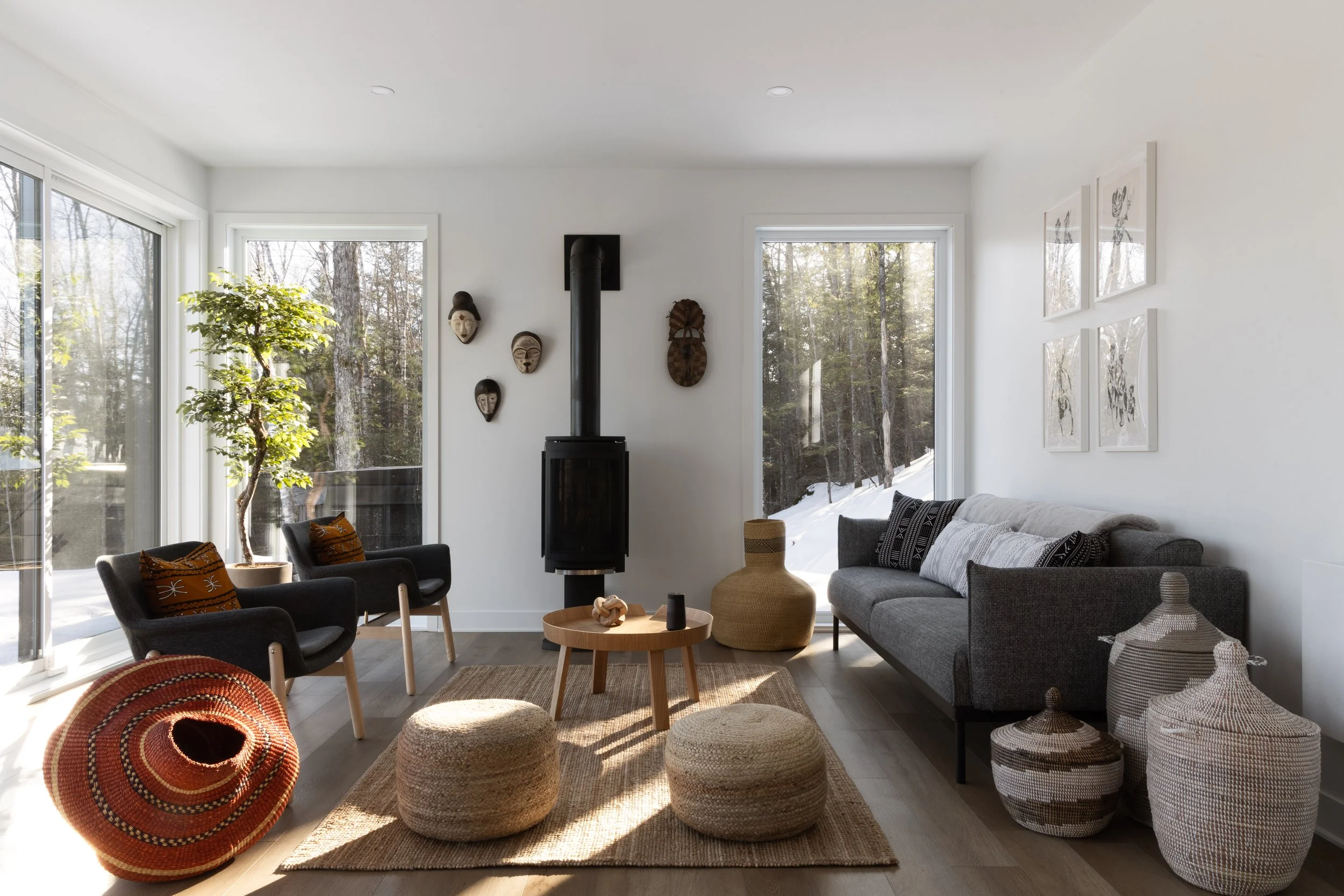 Bright living room with large windows, black wood stove, gray sofa, black armchairs with orange pillows, woven ottomans, and decorative African masks on the wall.