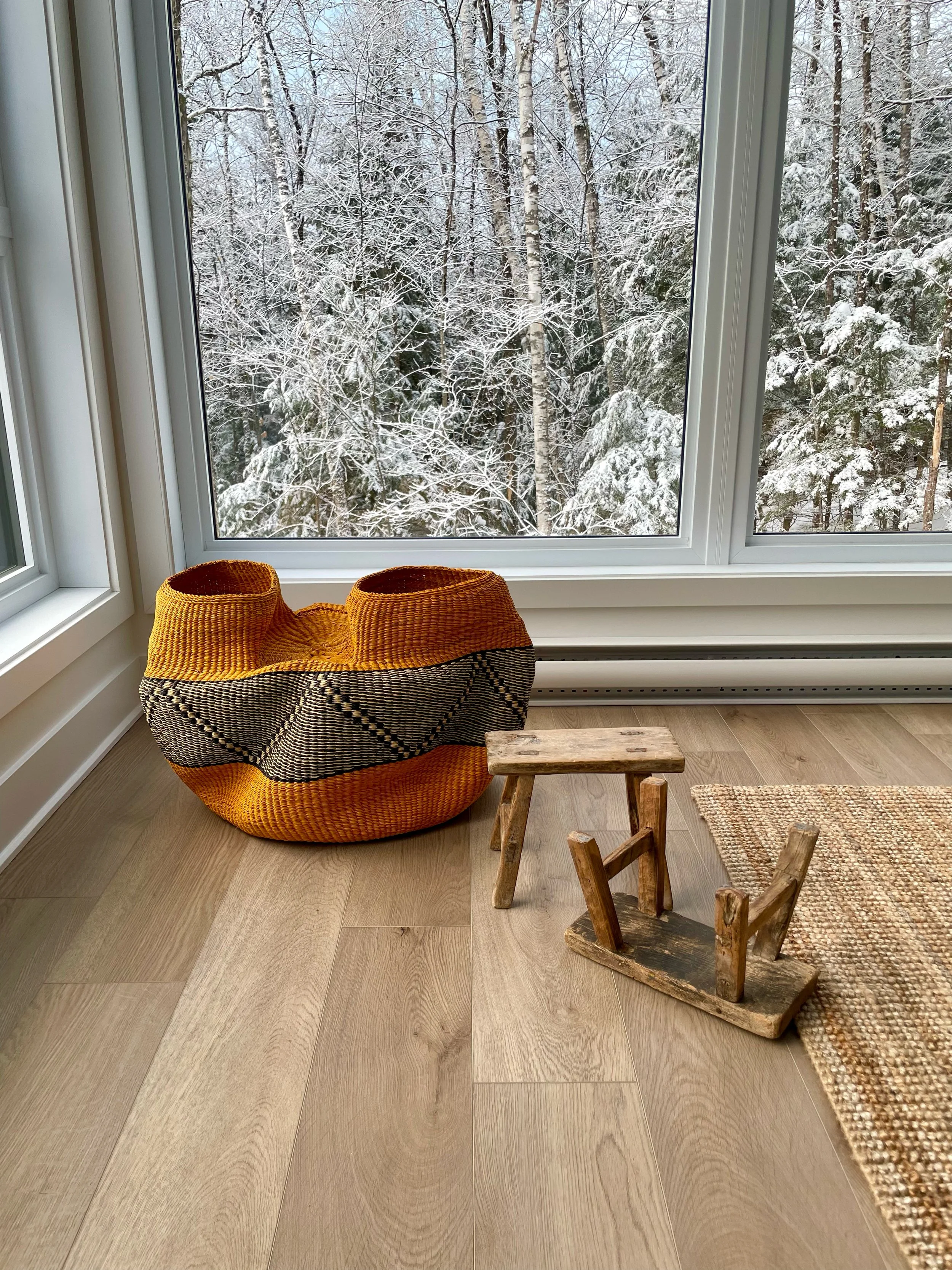 A room with wooden floor, a woven orange and black basket, a small wooden stool, a small wooden rocking chair, and part of a woven rug, with a large window showing snow-covered trees outside.