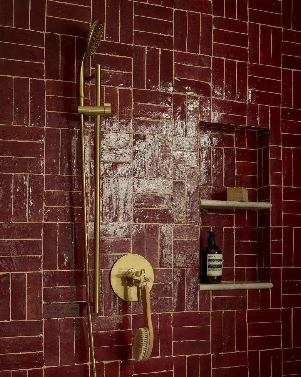 Shower with red brick zellige wall tiles in basketweave pattern, a brass showerhead and soap niche with a bottle of soap or lotion.