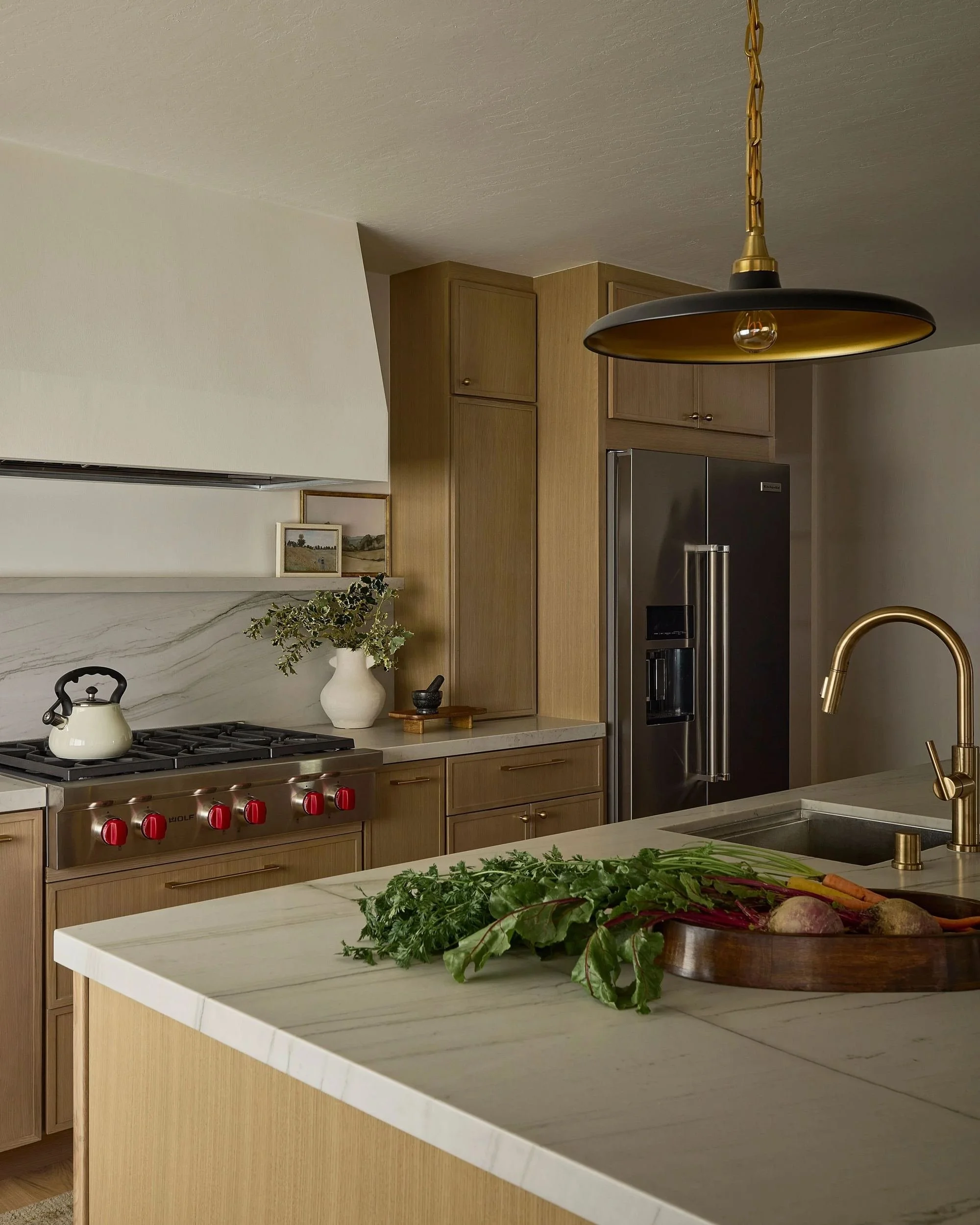 Kitchen with wooden cabinets, stainless steel refrigerator, black and gold pendant light, marble countertop, and vegetable arrangement in a bowl on the island.
