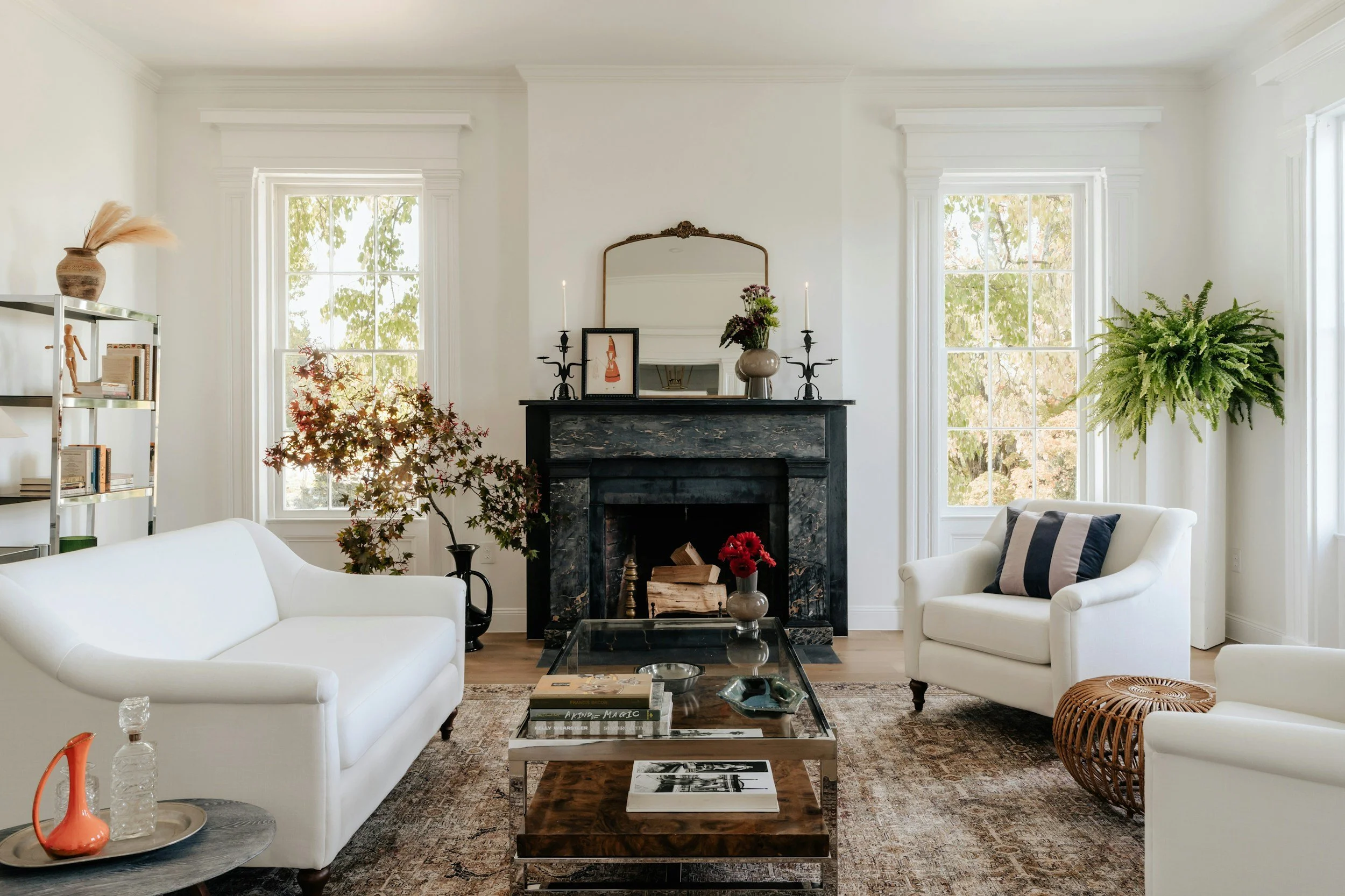 Living room with white sofas, a black fireplace, large windows with leafy views, and various decorative items including plants, candles, and books.