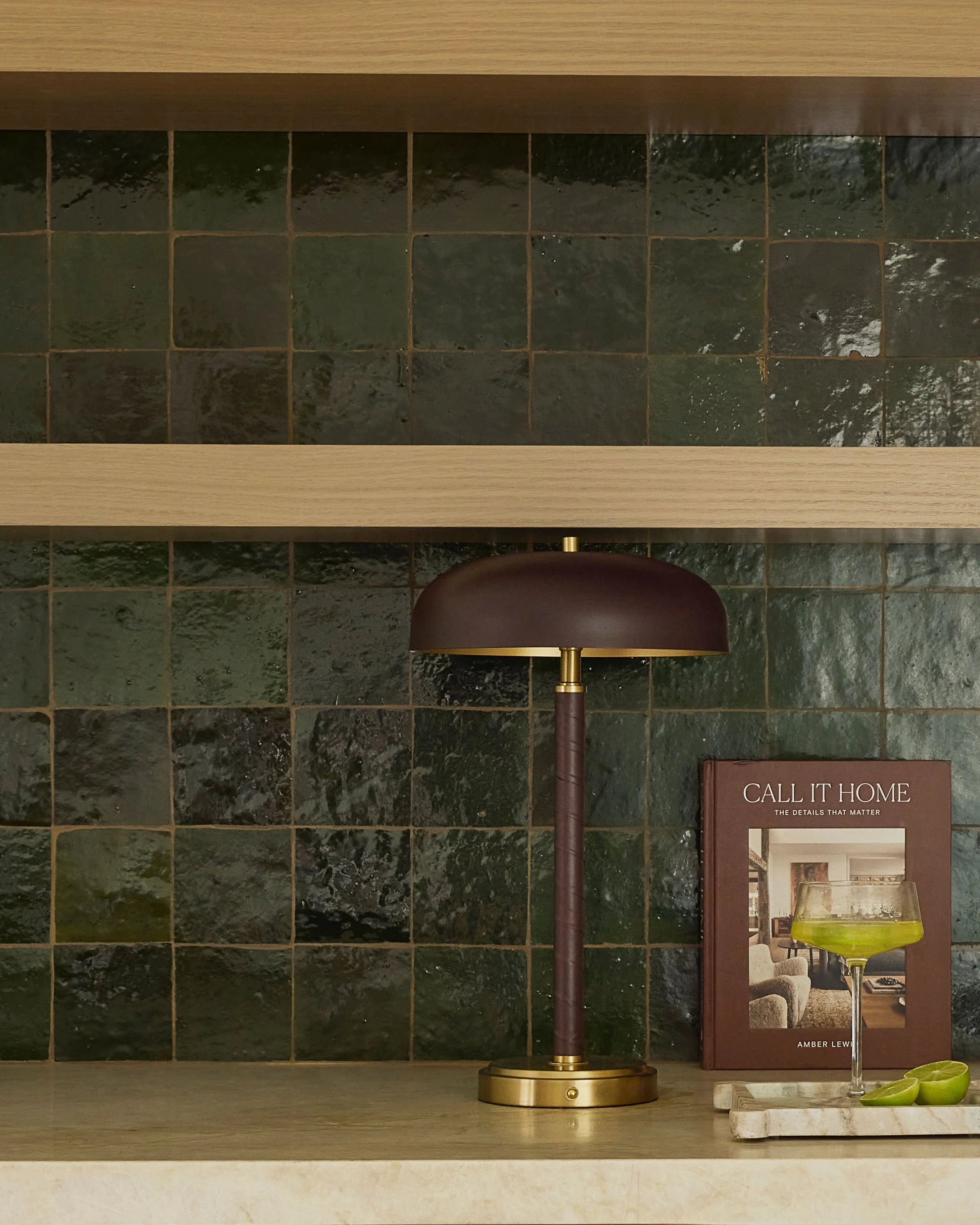 A decorative table lamp with a bronze base and dark brown shade, a book titled "Call It Home" with a partially visible arching green glass sculpture next to it, situated on a marble tray with small green bowls, all set against a black granite tile wa