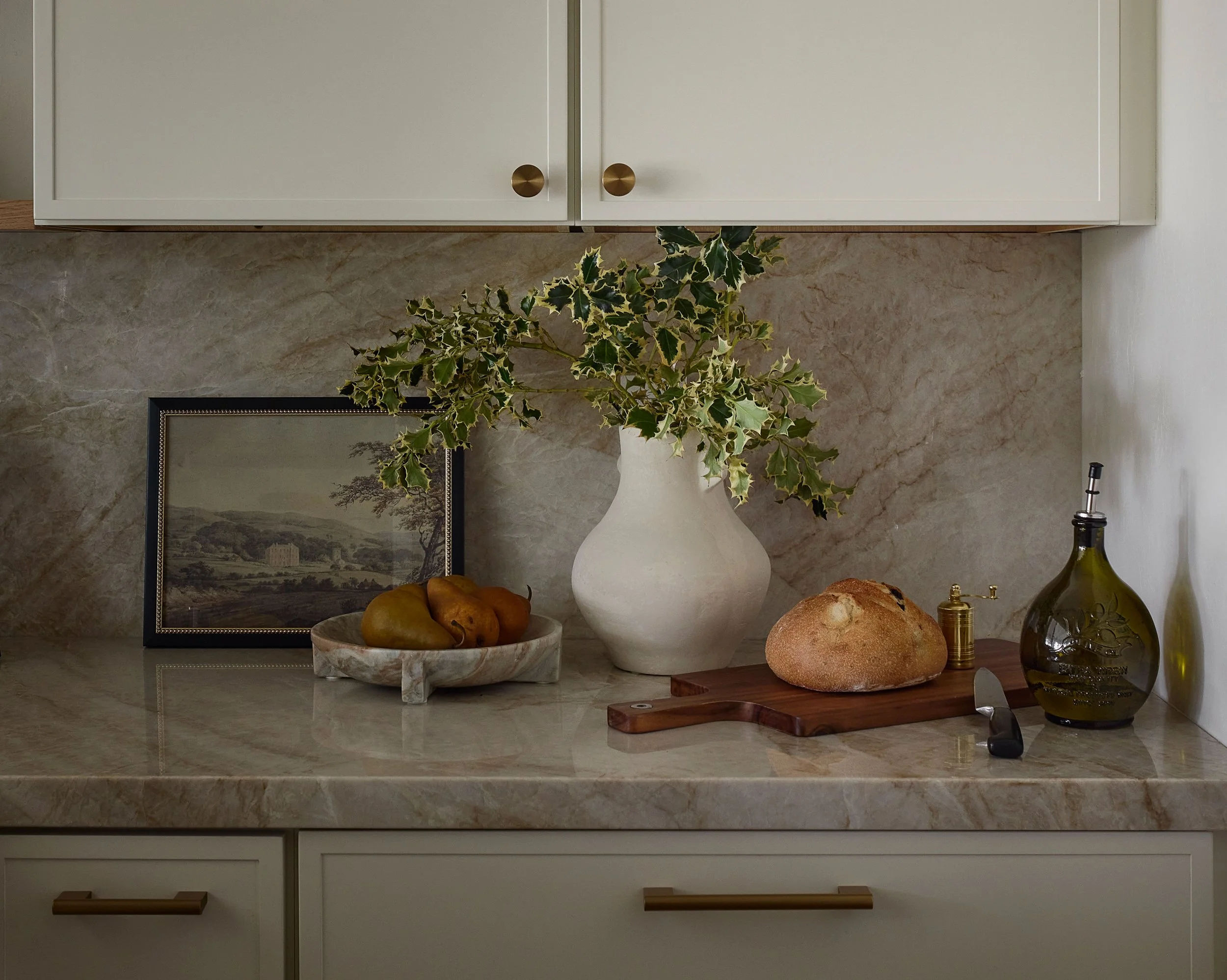 Kitchen countertop with a white vase holding faux holly leaves, a framed picture of a landscape, and bread rolls with a knife and oil bottles.