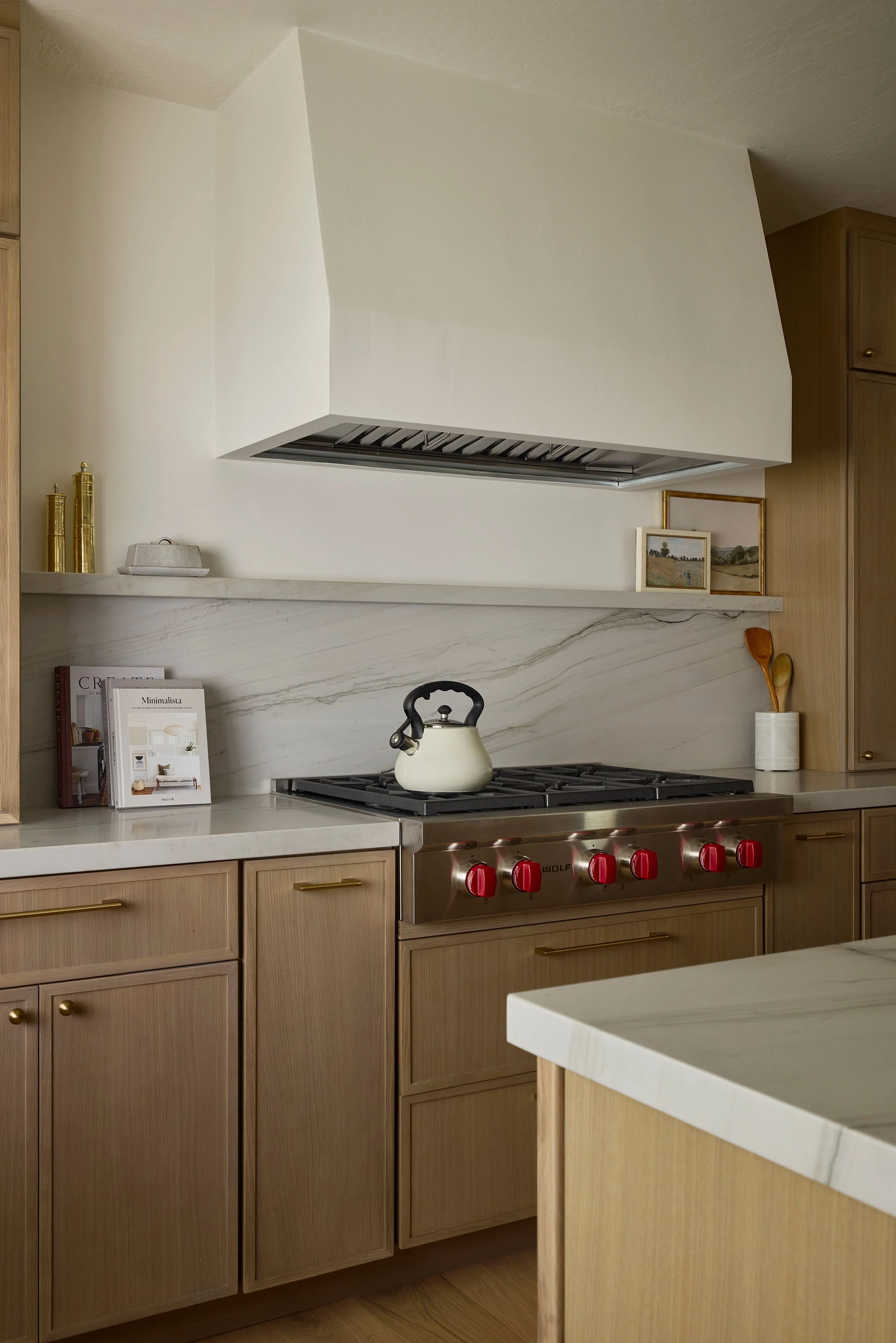 Kitchen with light wood cabinetry, white marble countertops, a stove with red knobs, a teapot on the stove, decorative framed pictures on the shelf, and kitchen utensils in a white container.