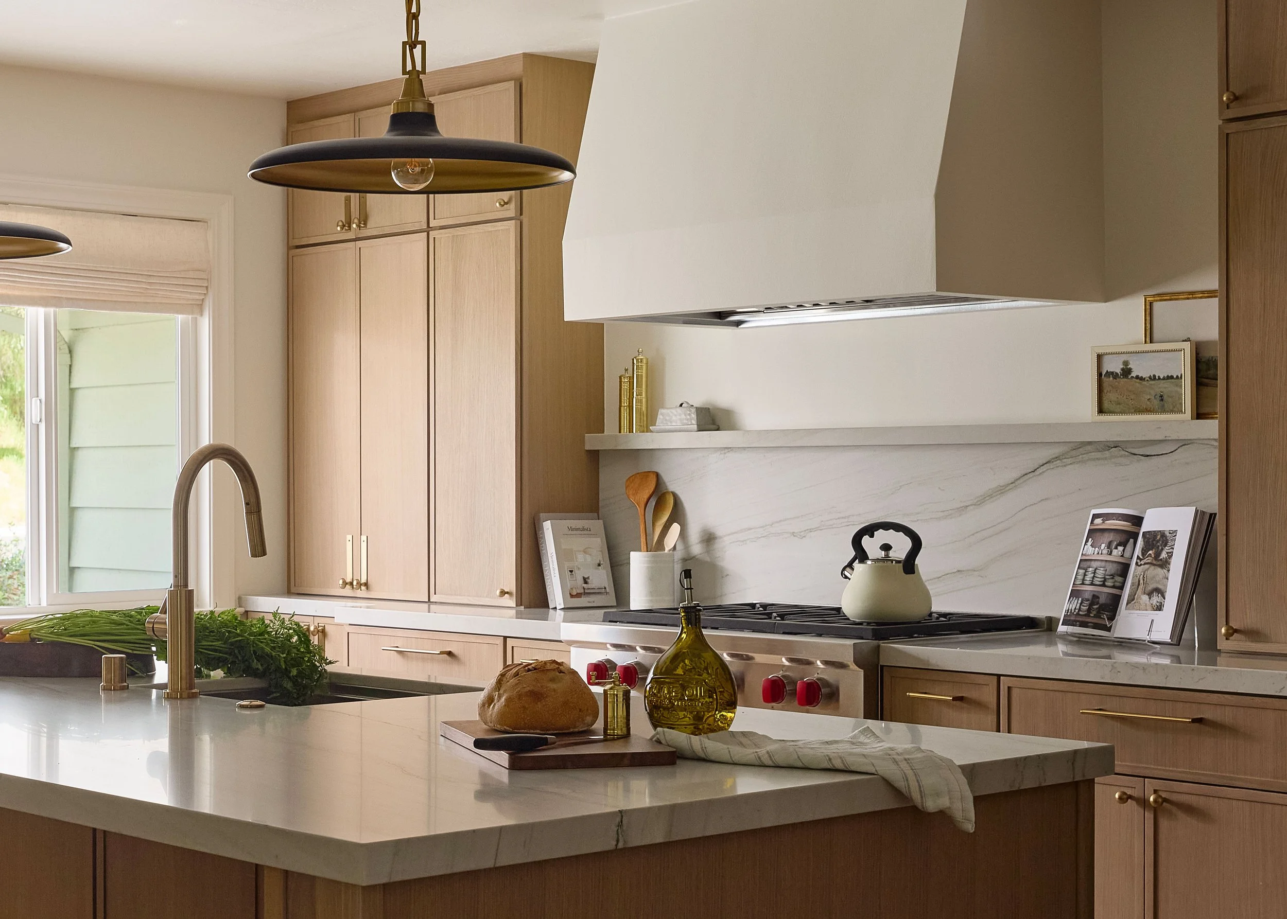 A modern kitchen with light wood cabinets, a marble backsplash, and a white marble island countertop. There are various kitchen items including a loaf of bread, bottles of olive oil, and framed pictures on a ledge.