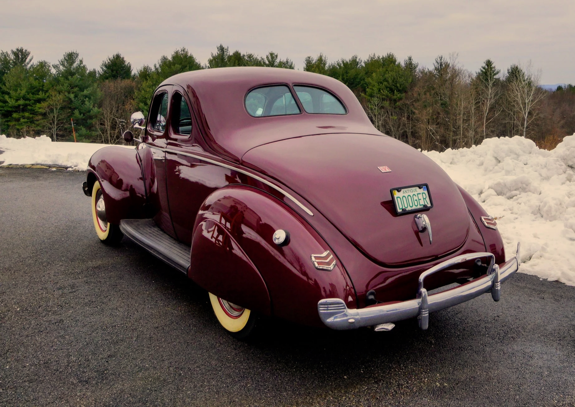 A vintage maroon-colored car with a rounded body parked on an asphalt road with snow piles on the side, a forested area with trees in the background, and a cloudy sky.