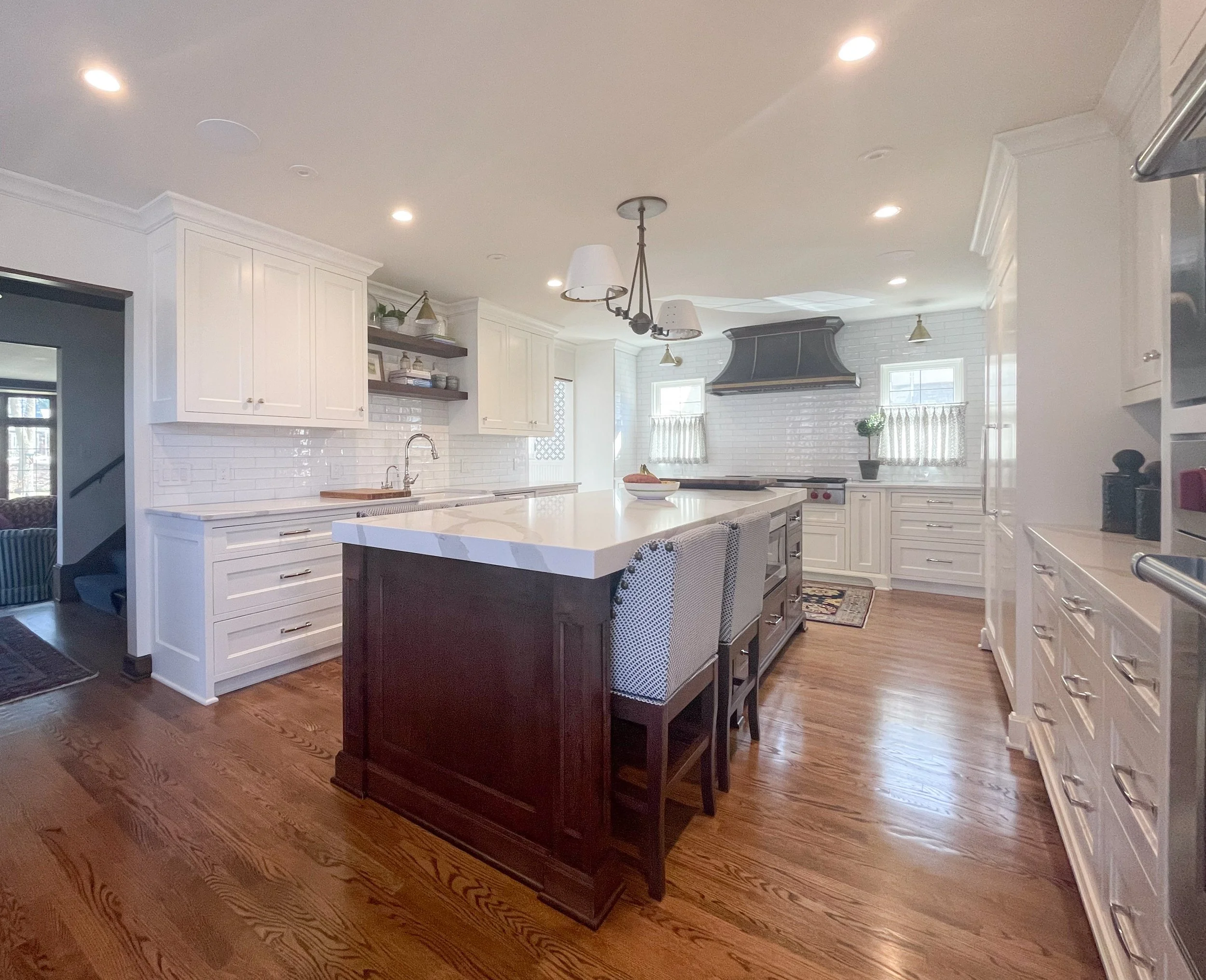 Bright kitchen with white cabinets, a central island with a dark wood base and white countertop, hardwood floors, and natural light coming through two windows with curtains. There are open shelves, a range hood, and seating at the island.