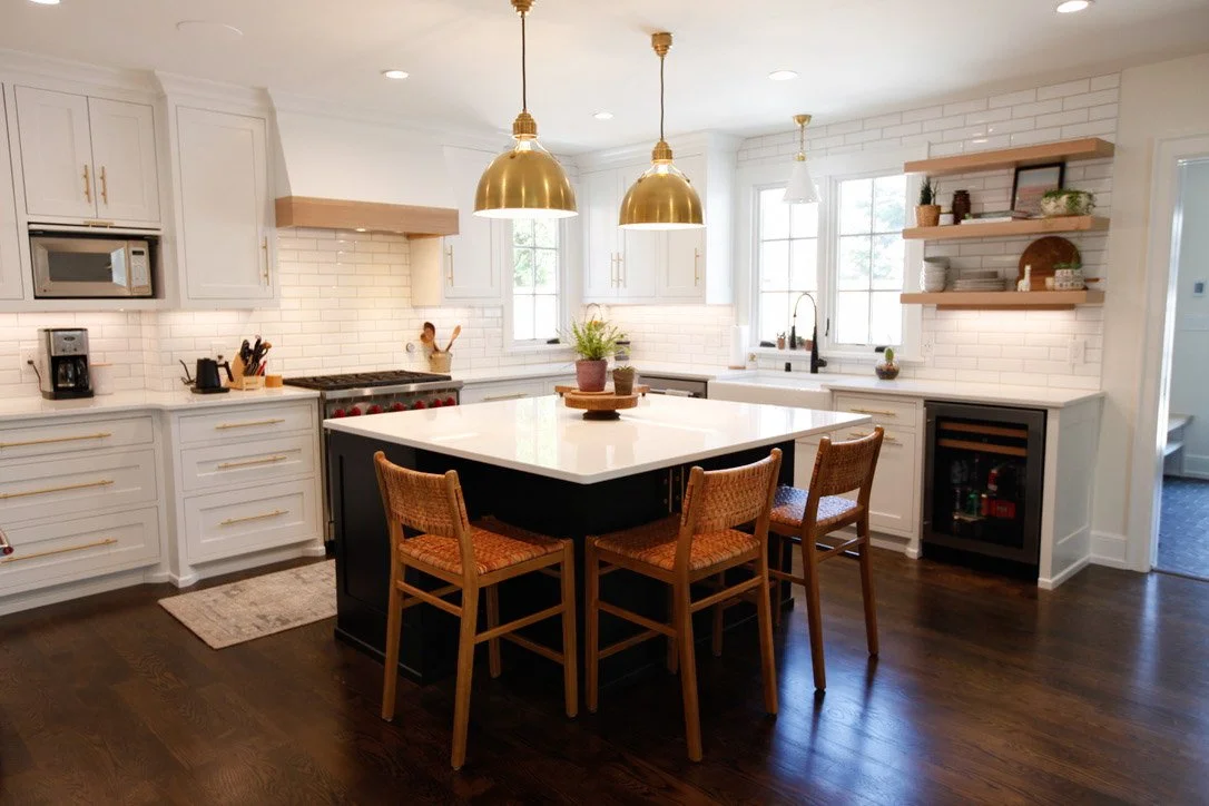 Bright kitchen with white cabinets, a black island with a white countertop, hanging gold pendant lights, open shelving, and a window with natural light.