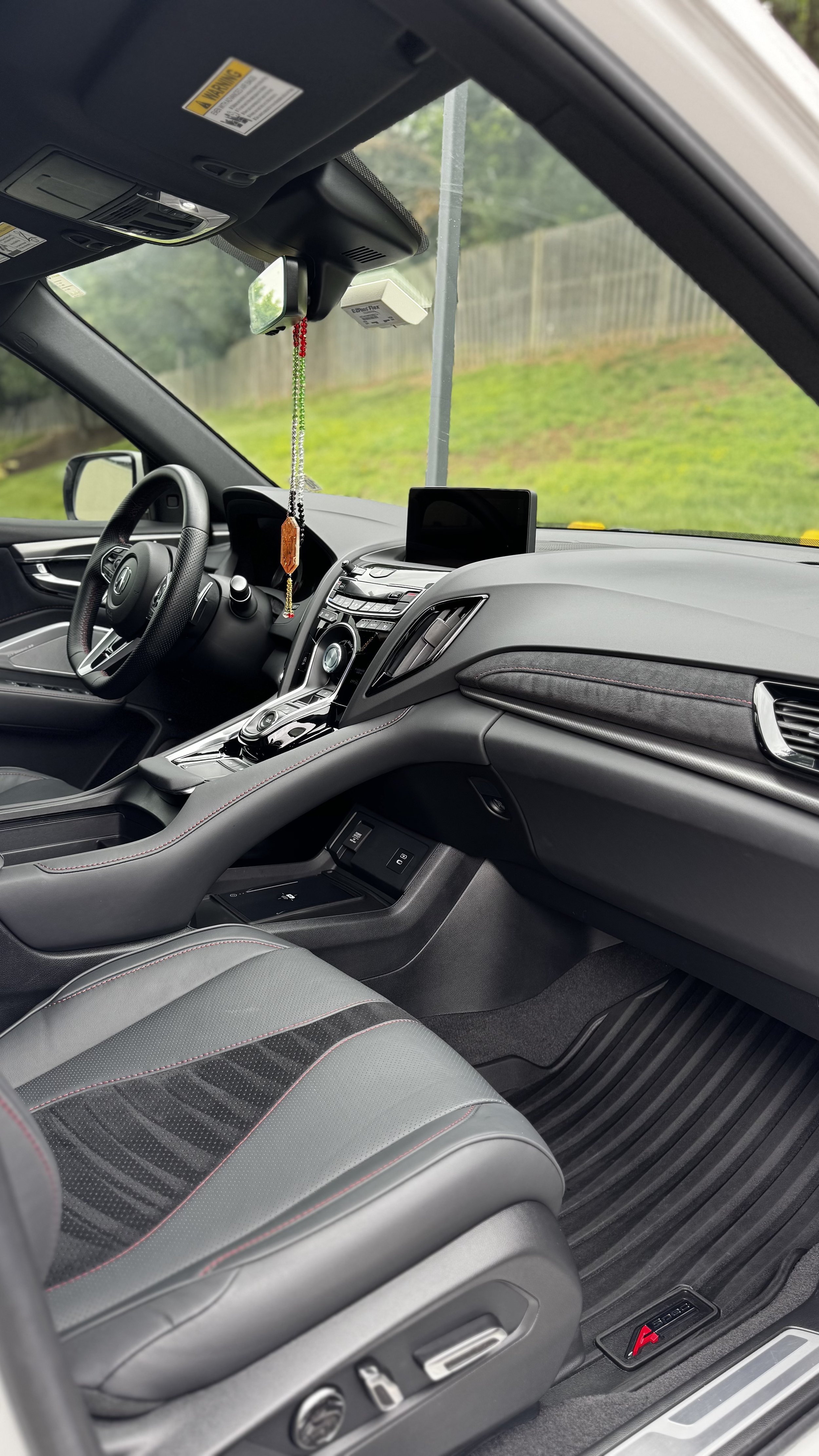 Interior of a modern car showing the driver's seat, steering wheel, dashboard, and center console. The dashboard has a digital display screen, and the seat has red stitching accents. The car is parked outdoors with a grassy hill and wooden fence visi