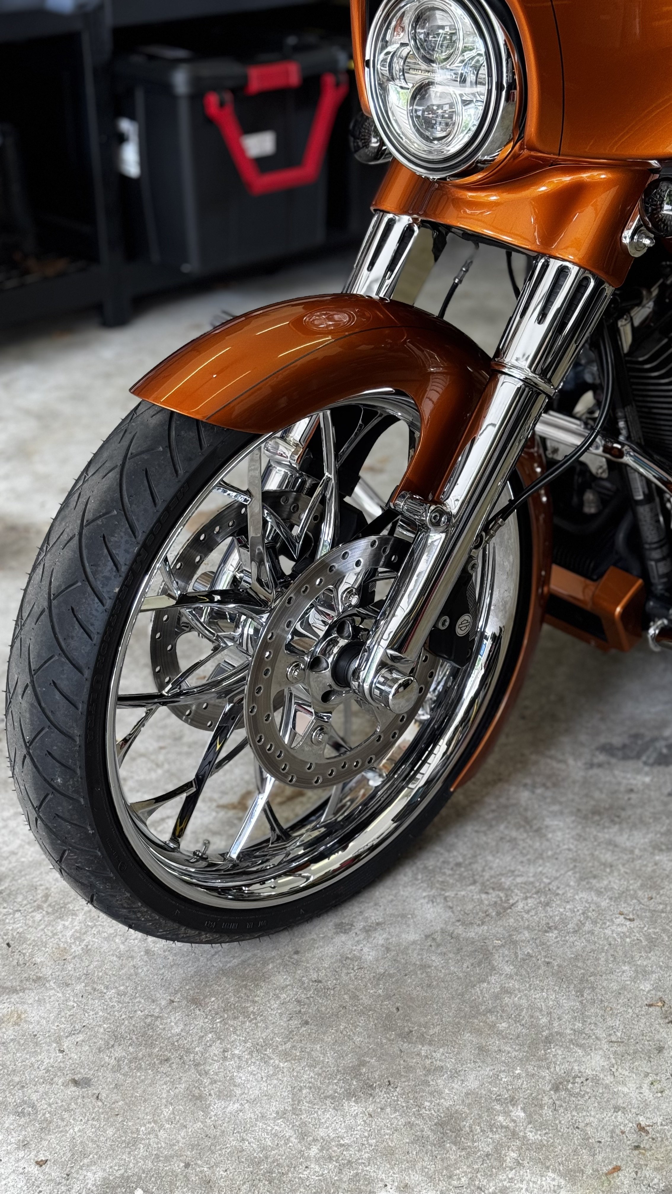Close-up of a motorcycle front wheel with chrome spokes, disc brake, and orange front fender, parked on a concrete surface.