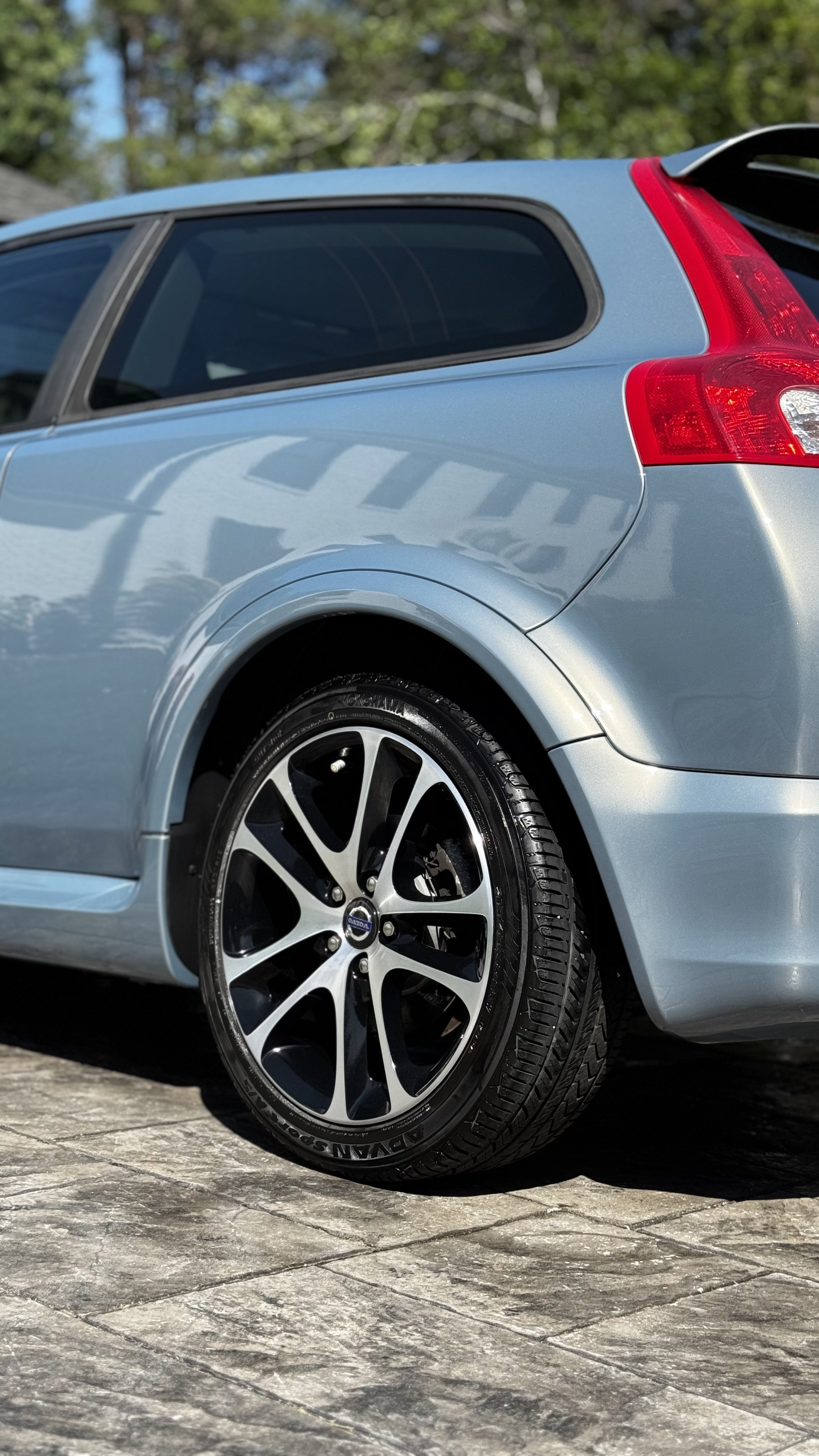 Close-up of the rear end of a silver car showing the wheel, tire, rear taillight, and part of the rear window, parked on a stone-paved driveway with green trees in the background.