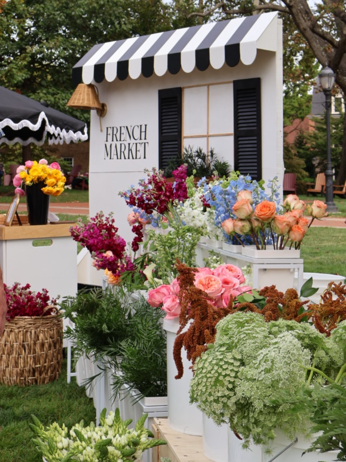 A flower stand at a French Market, with colorful flowers including roses, hydrangeas, and other blooms in white pots and baskets, in an outdoor garden setting.