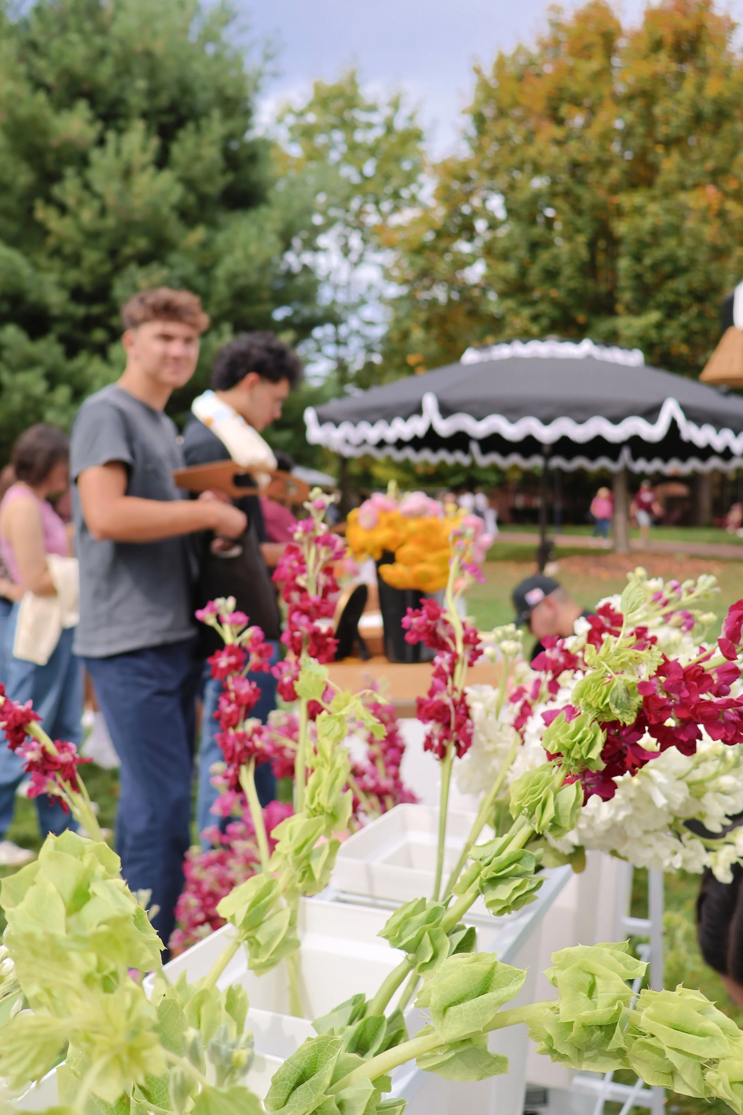 Close-up of pink, white, and green flowers on a table at an outdoor event, with people in the background near a black canopy with white trim, green trees, and a woman wearing a pink shirt.