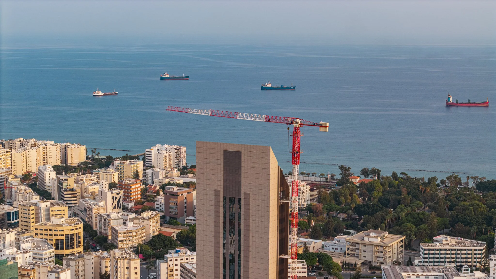 Cityscape with tall buildings and a construction crane overlooking the ocean with several ships sailing in the water.