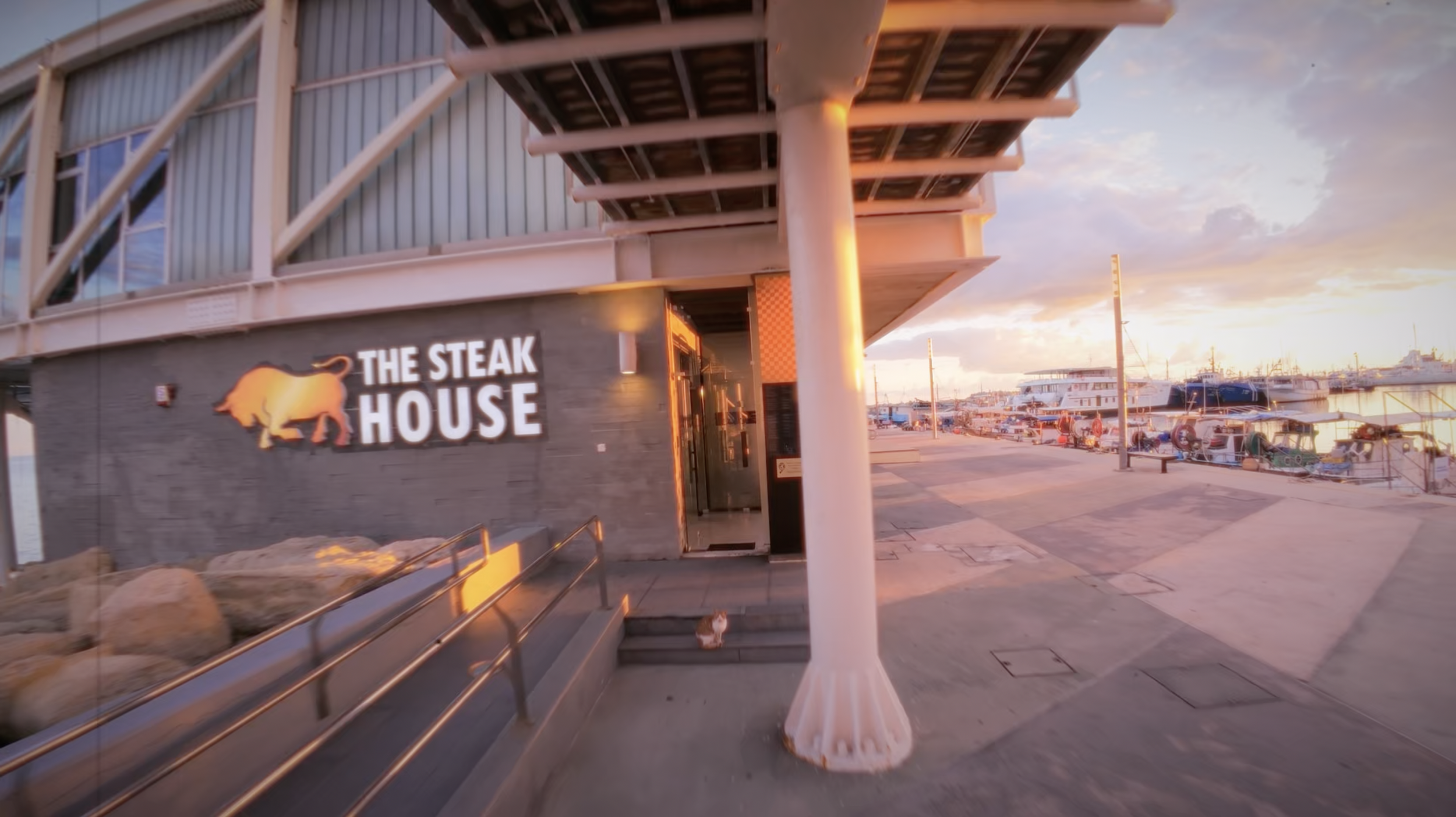 Exterior shot of The Steak House in Limassol during golden hour, showing the entrance, signage, and marina walkway captured from an FPV approach.