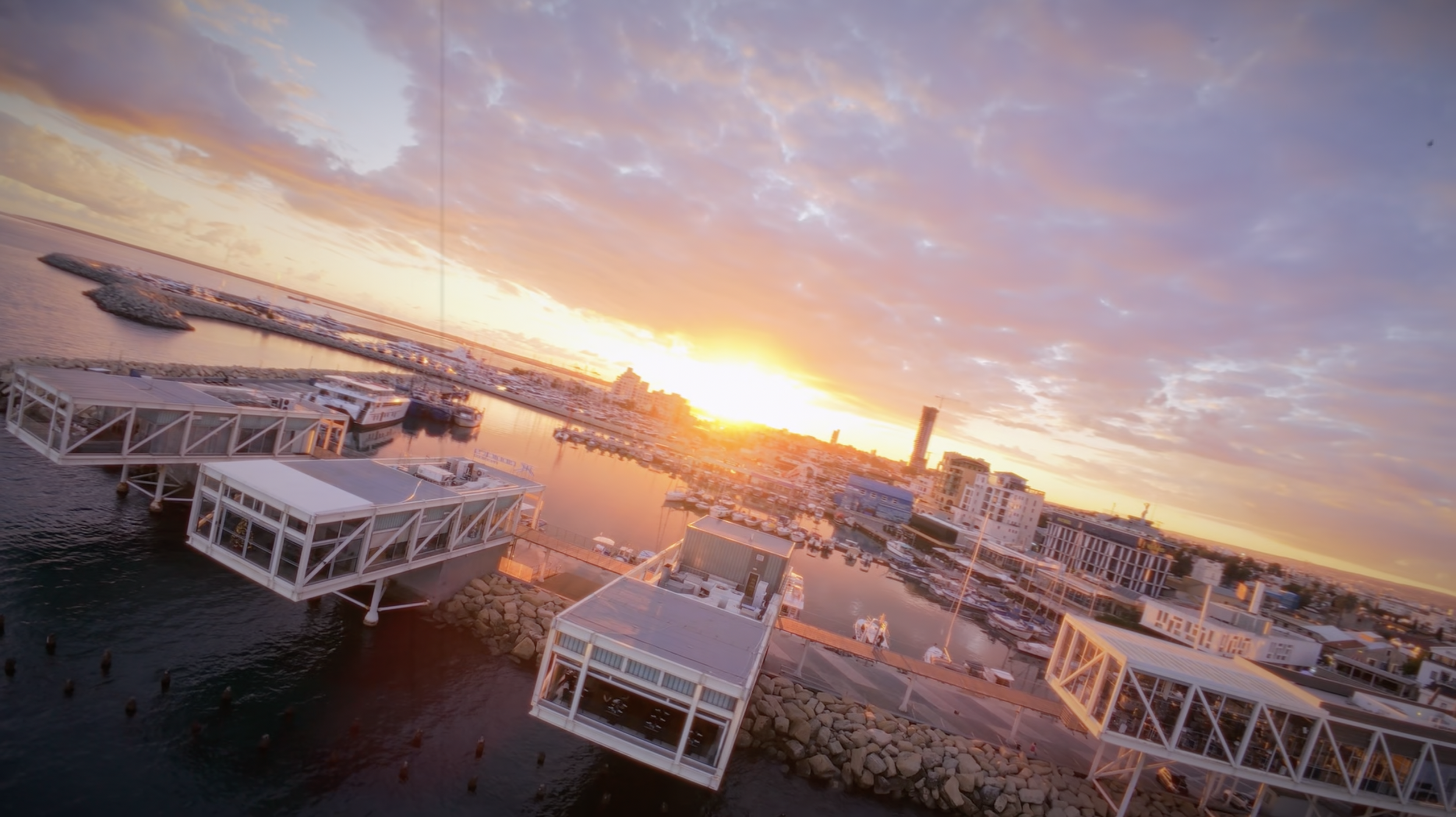 Aerial sunset view over Limassol Marina with the floating restaurant platforms, boats, and city skyline framed by warm evening light.