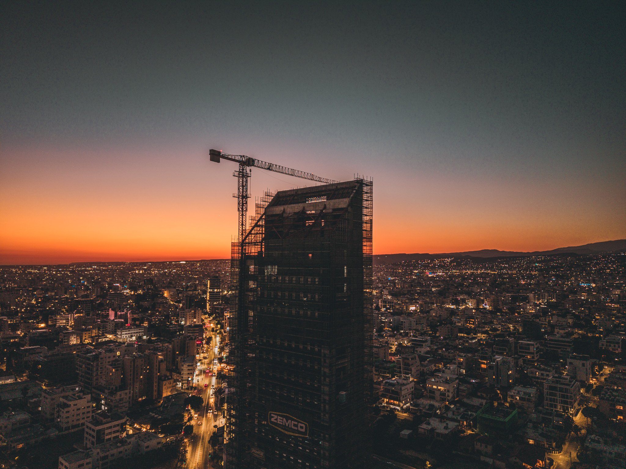 A cityscape at sunset with a tall building under construction and a crane on top, overlooking a city filled with lights.