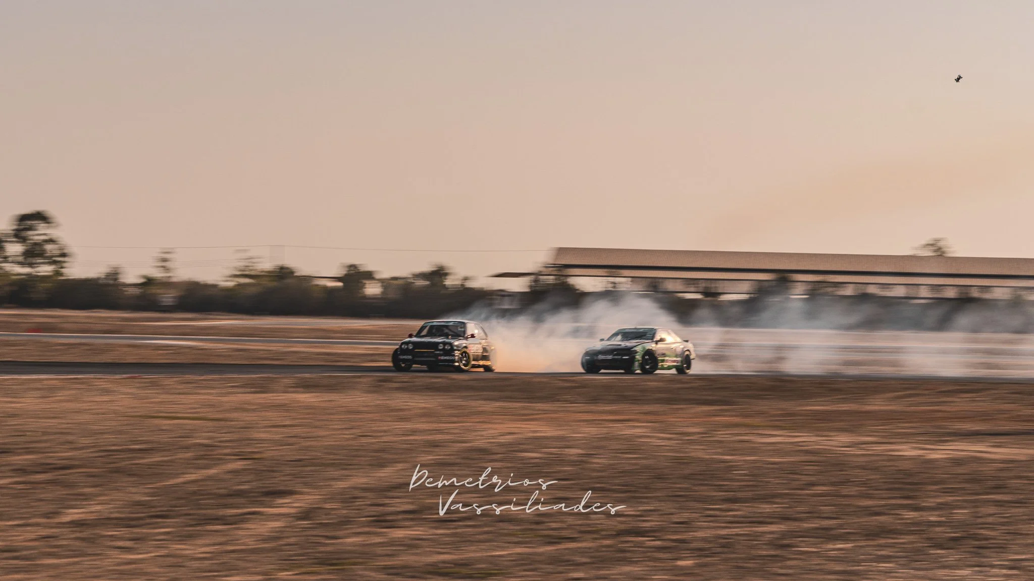 Two drift cars entering a wide corner at sunset with smoke trails.