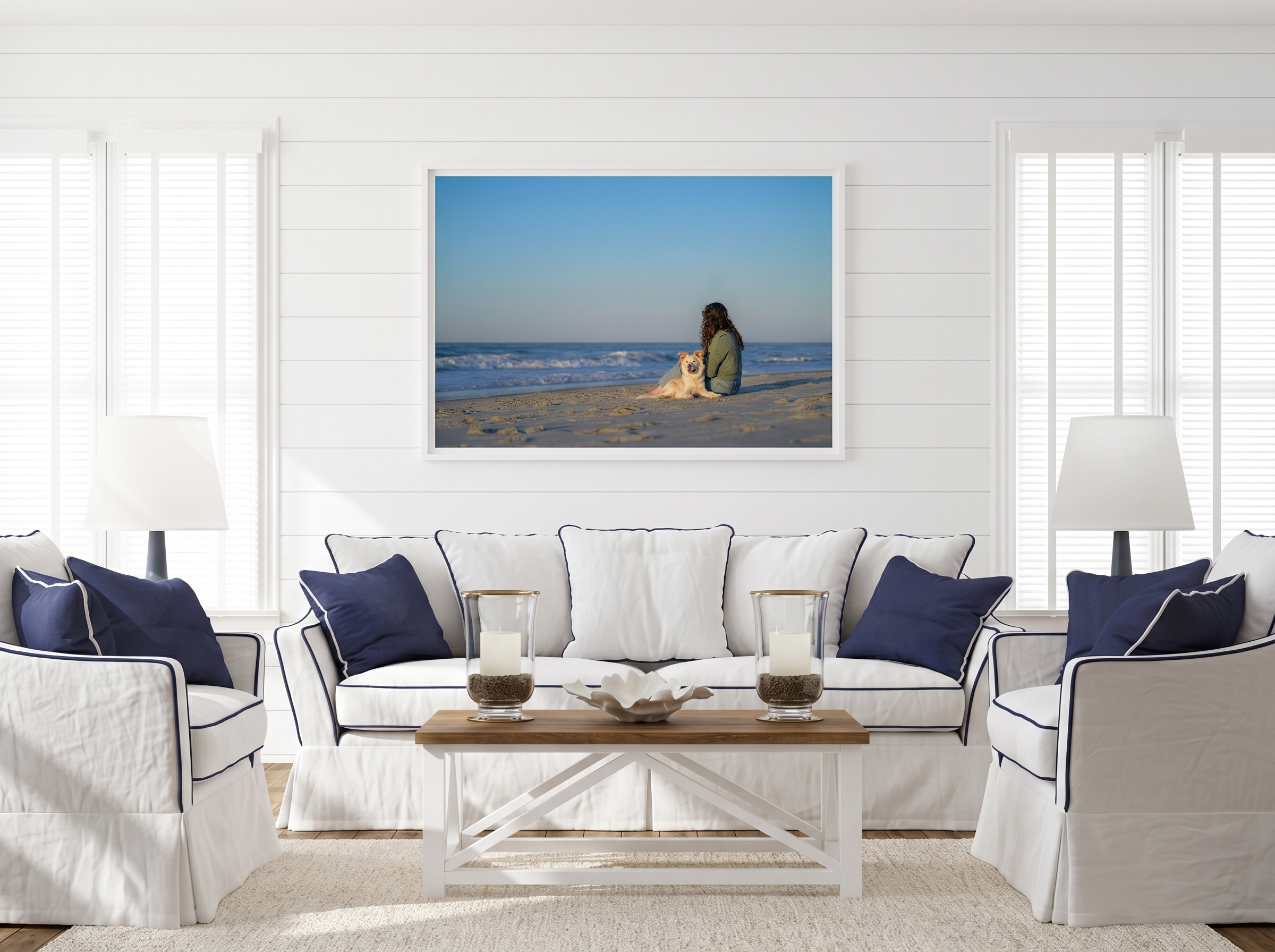 A large framed canine portrait hangs above a white coastal sofa — a woman and her cream-colored dog sit together on a sandy beach facing the ocean, photographed by Jennifer Wakefield Photography on the Crystal Coast of North Carolina.