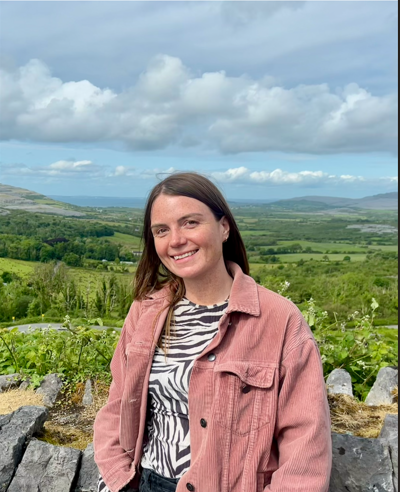 A woman smiling outdoors with rolling green hills, vineyards, and a cloudy sky in the background.