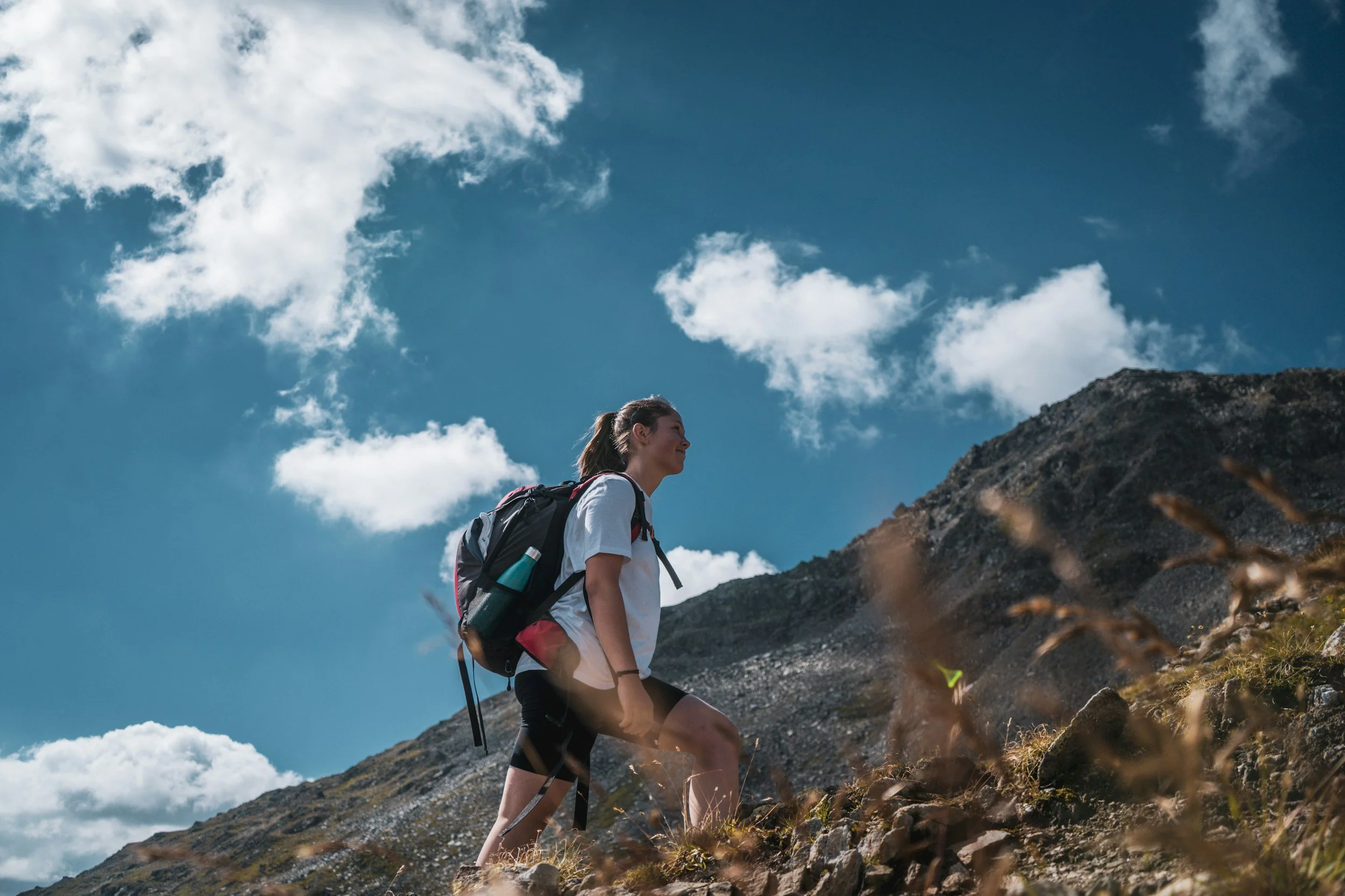 A woman hiking up a mountain trail with a backpack under a partly cloudy sky. 