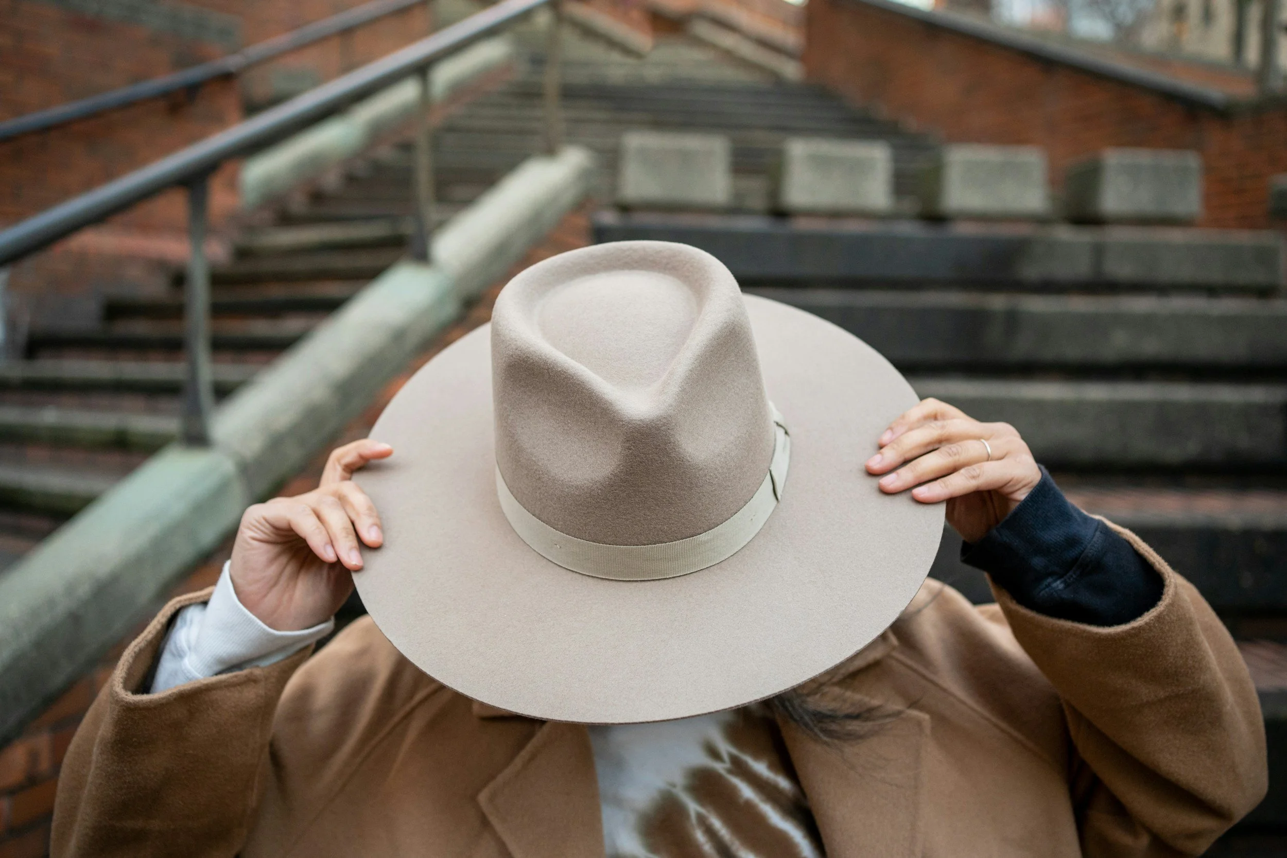 Person adjusting a beige wide-brimmed hat with a band, standing on outdoor stairs with a brick building background.