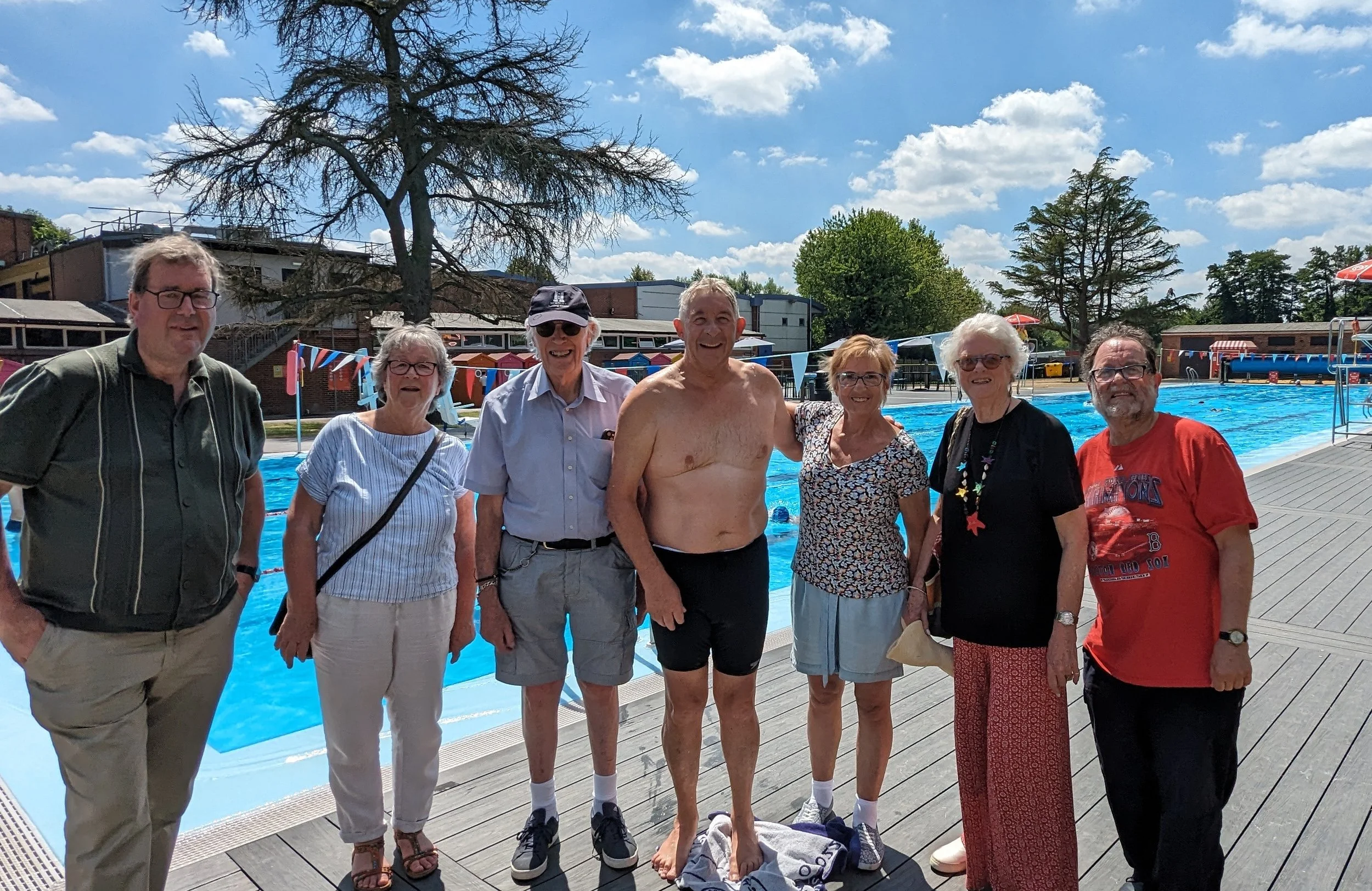 Group of seven people standing by an outdoor swimming pool on a sunny day, some wearing casual clothes and others in swimwear, with a large tree and buildings in the background.
