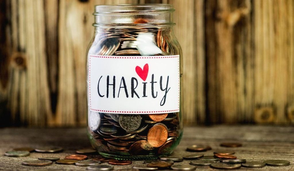 A glass jar filled with coins, placed on a wooden surface with some coins scattered around, and a label that says 'Charity' with a red heart.