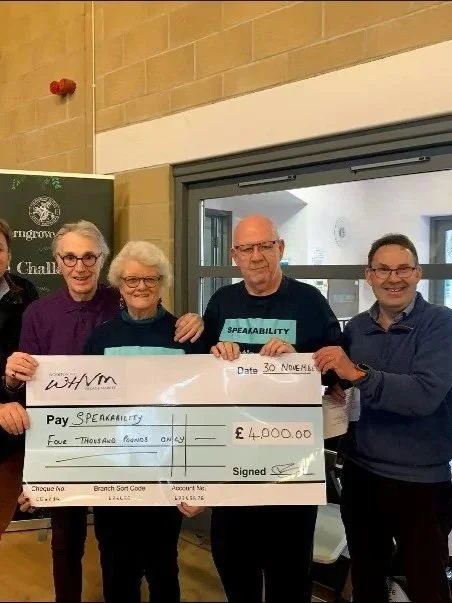 Four people standing indoors, holding a large check for £4,000 made out to Speakability, with a woman and a man holding the check on either side. They are smiling and dressed in casual attire.