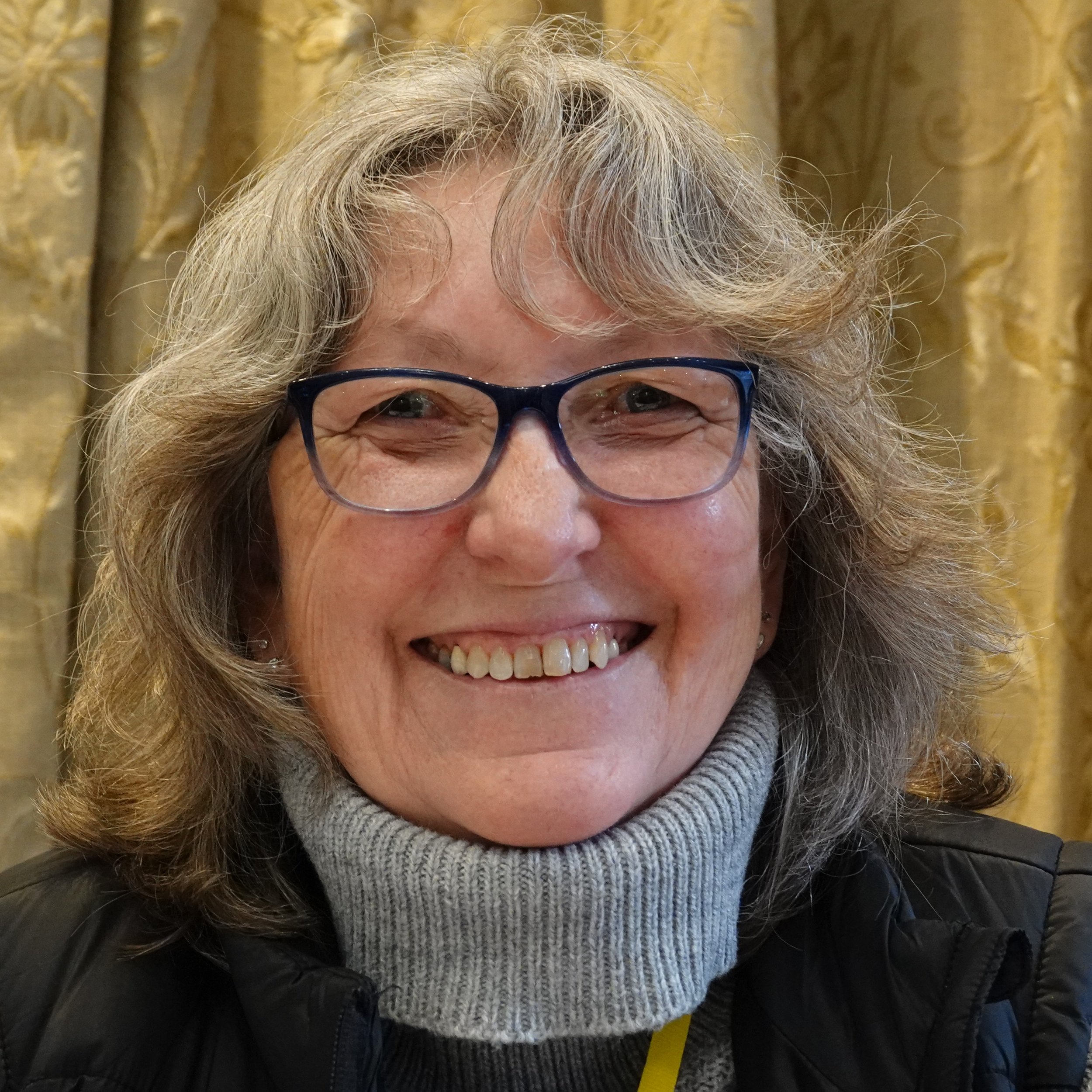 A woman with curly gray hair, glasses, and a happy smile, wearing a turtleneck sweater and a black jacket, with a gold curtain background.