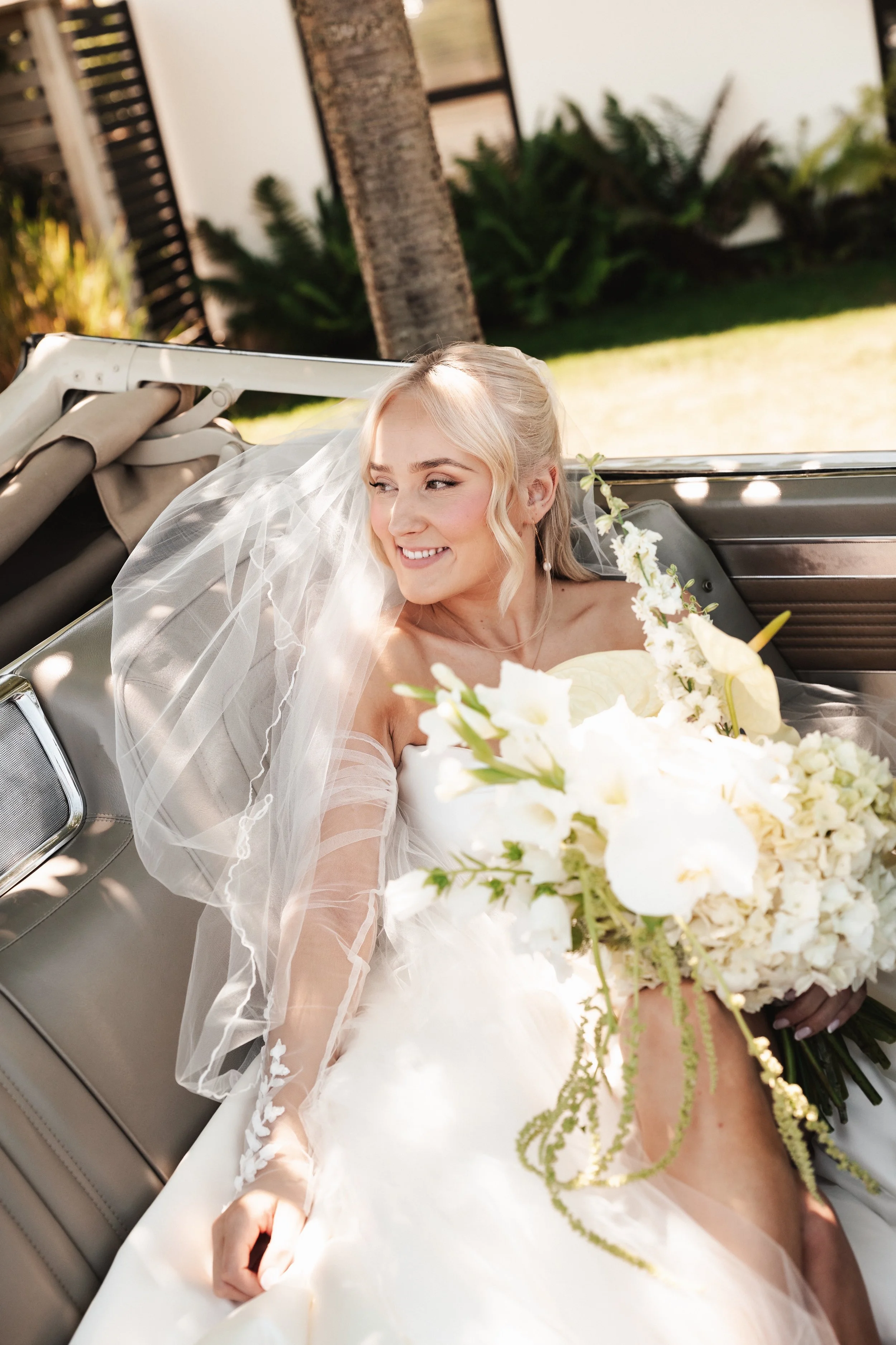 A smiling bride sitting in a convertible car holding a bouquet of white flowers, with a veil and wedding dress.