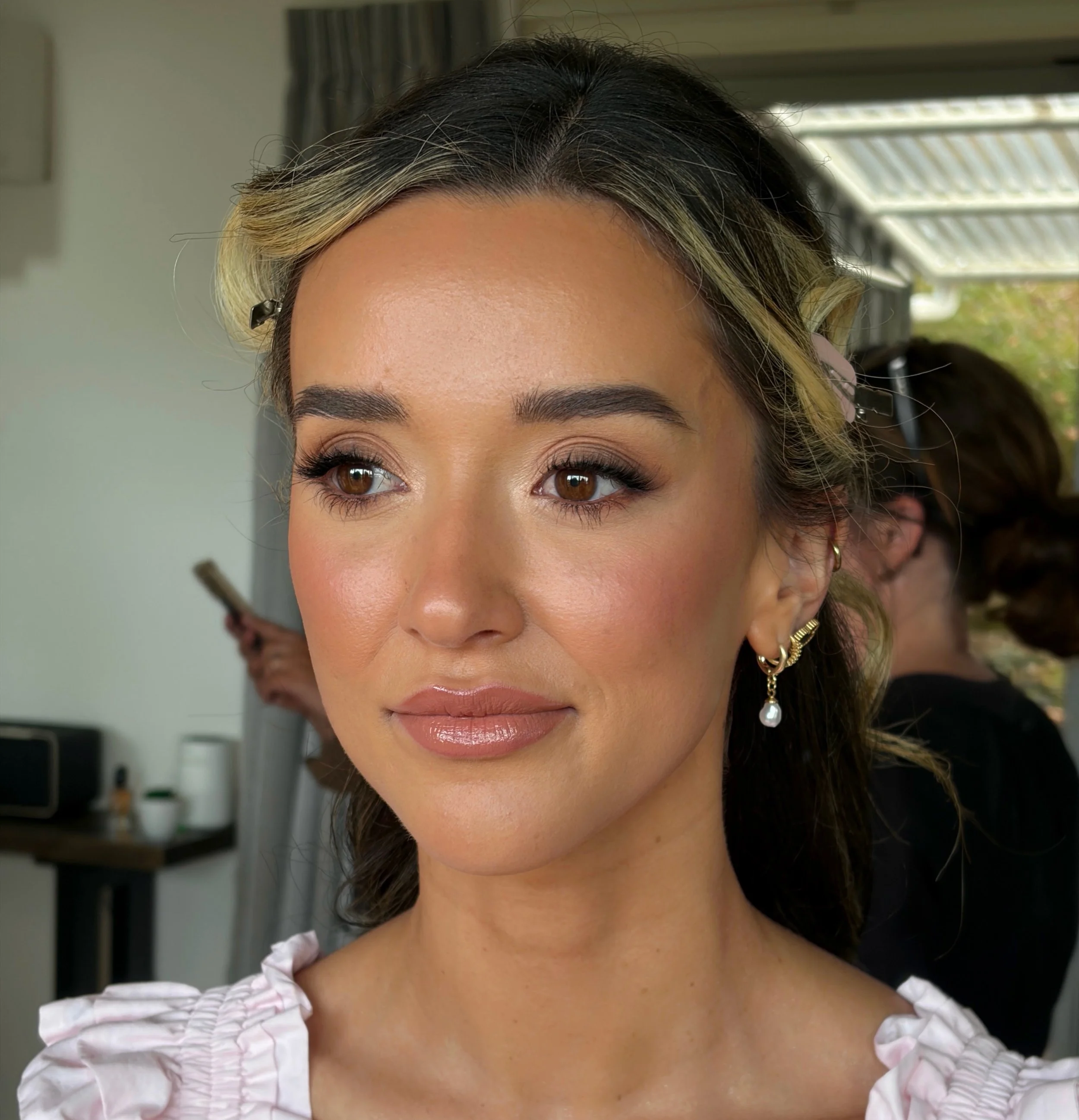 Close-up of a woman with dark brown and blonde hair wearing earrings and a white ruffled blouse, indoors with a window in the background.