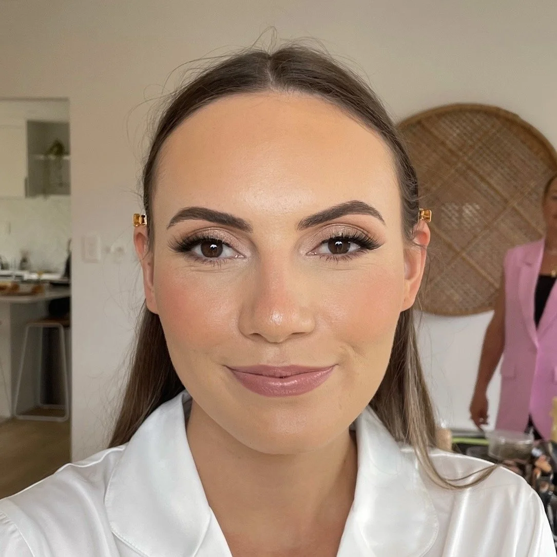 Close-up of a woman with brown hair, wearing makeup, smiling, in a white shirt, with a woman in pink in the background in a home setting.