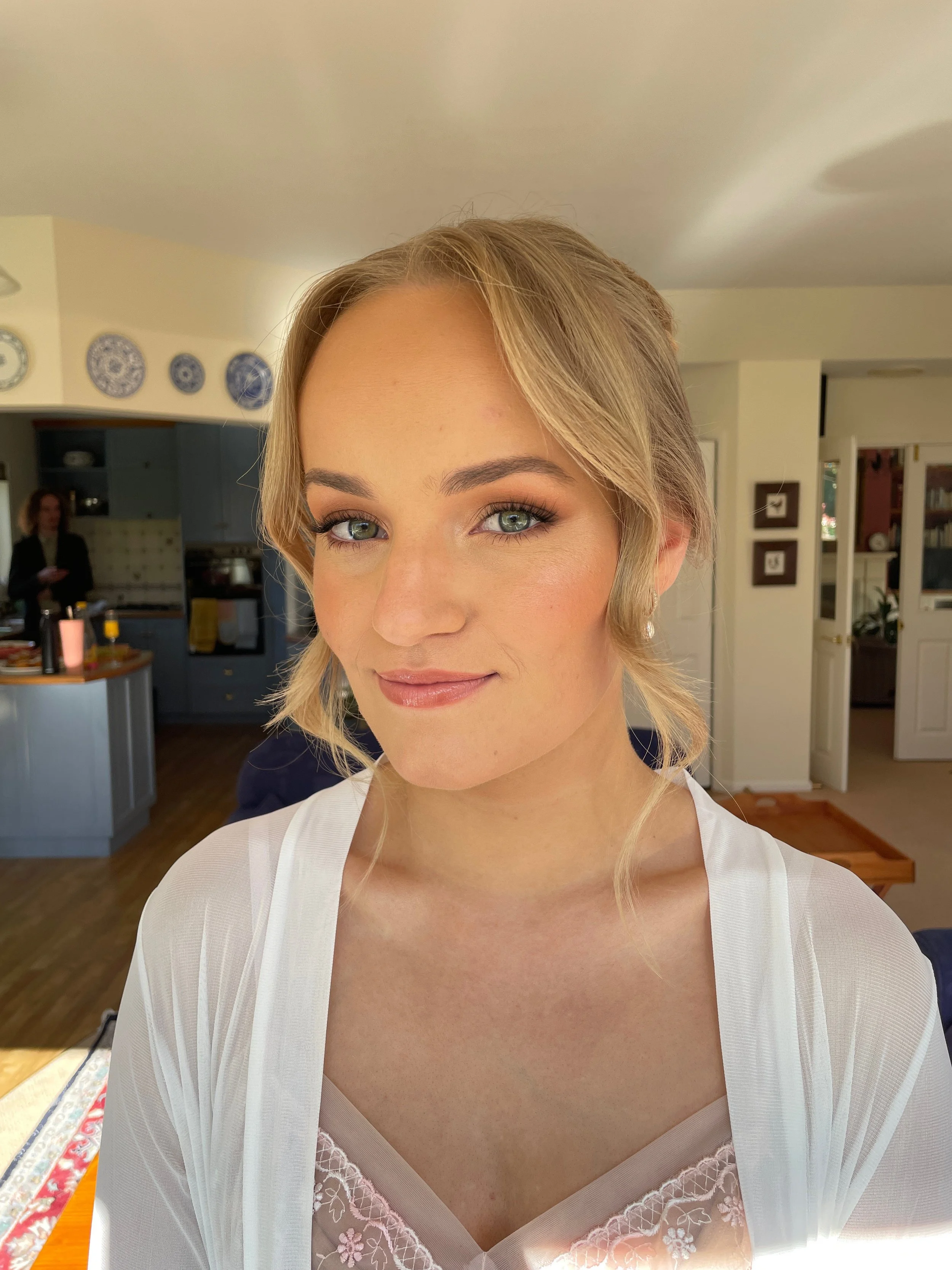 Close-up of a young woman with blonde hair and green eyes wearing a white sheer robe and pink lace lingerie, smiling softly inside a home with blue cabinets and decor on the walls.