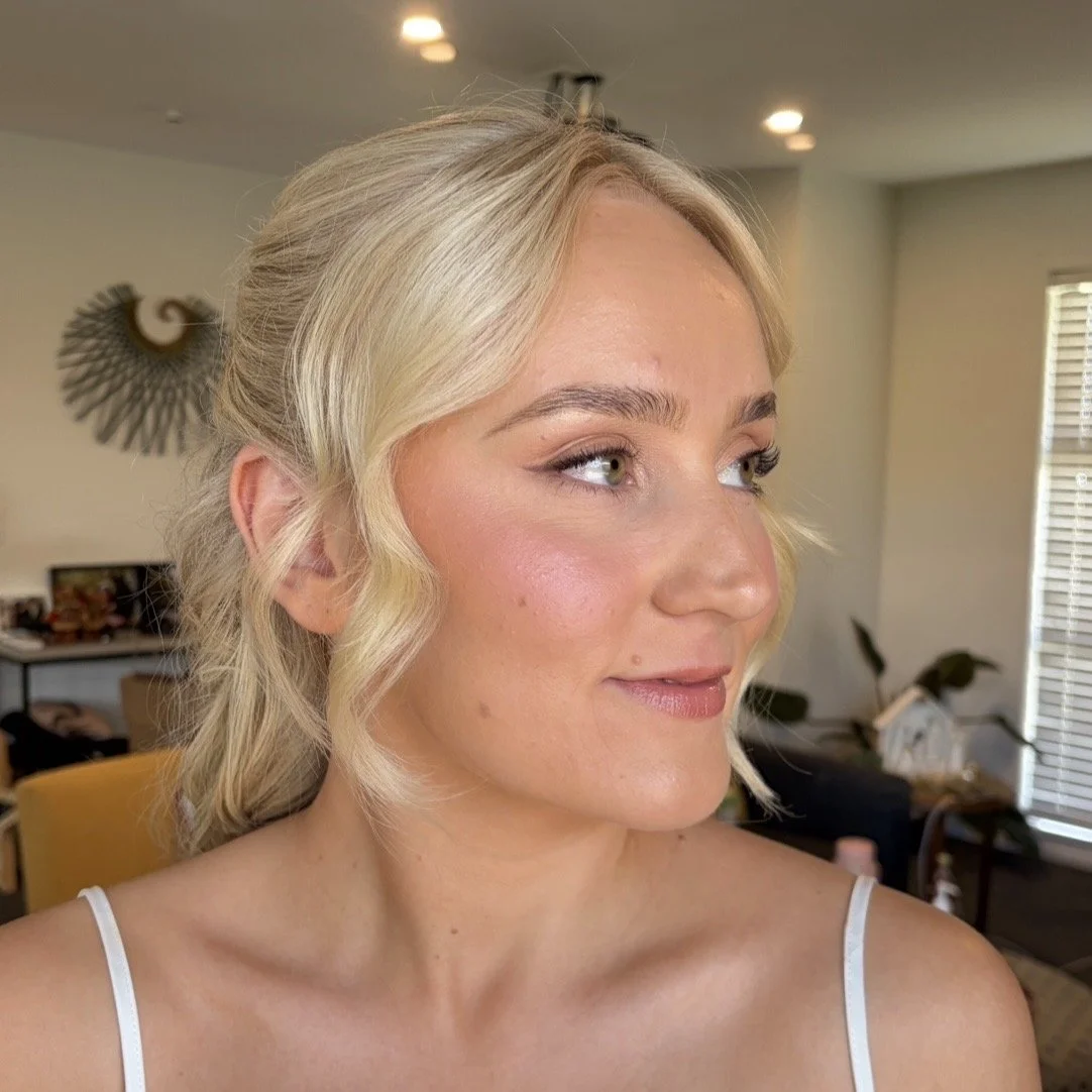 A blonde woman with wavy hair and light makeup, looking to the side, in a living room with artwork and furniture in the background.