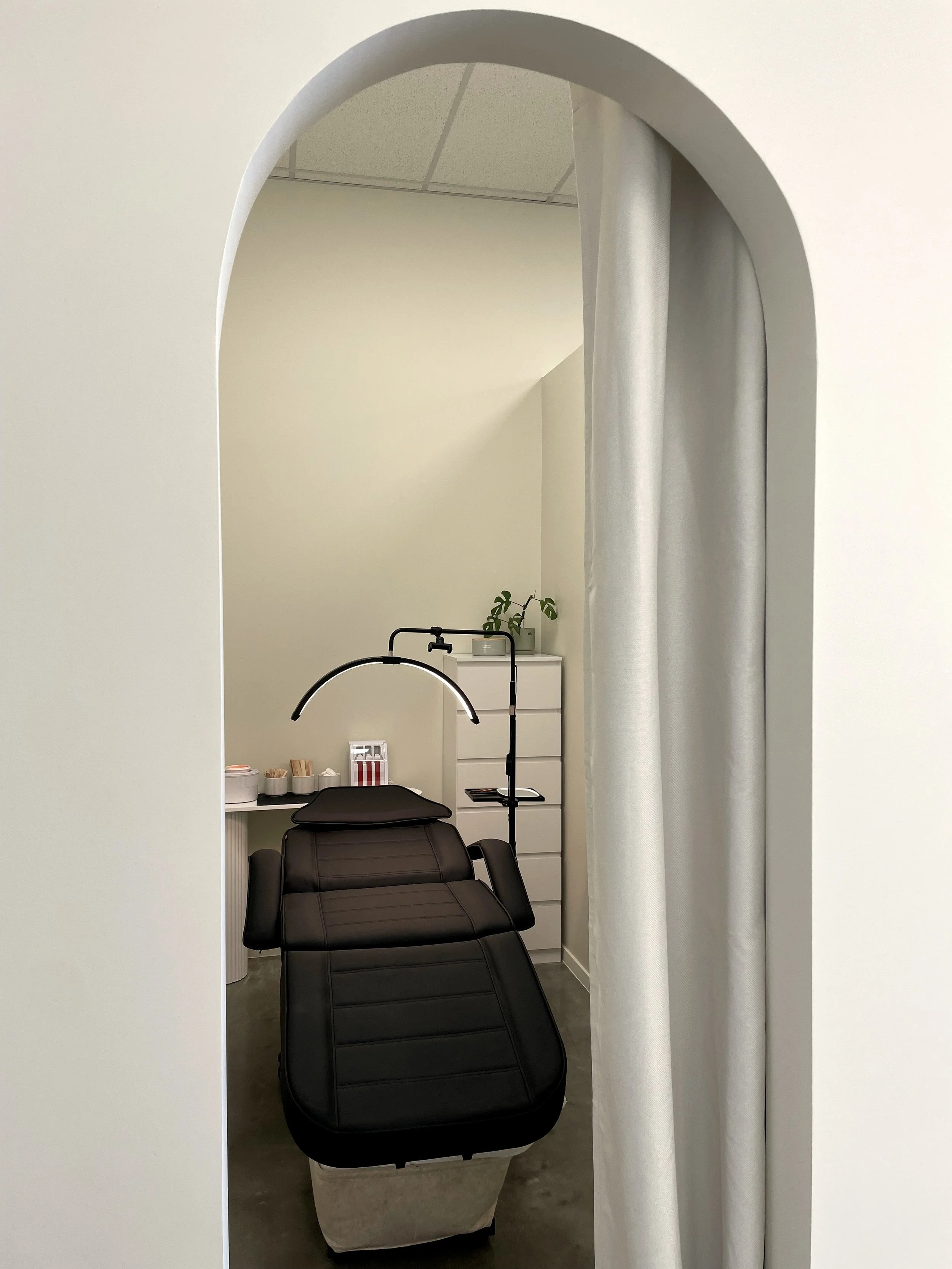 Medical or beauty treatment room with a black treatment chair, a lamp above it, and a white storage unit with plants and supplies in the background, view through an arched doorway with a curtain.