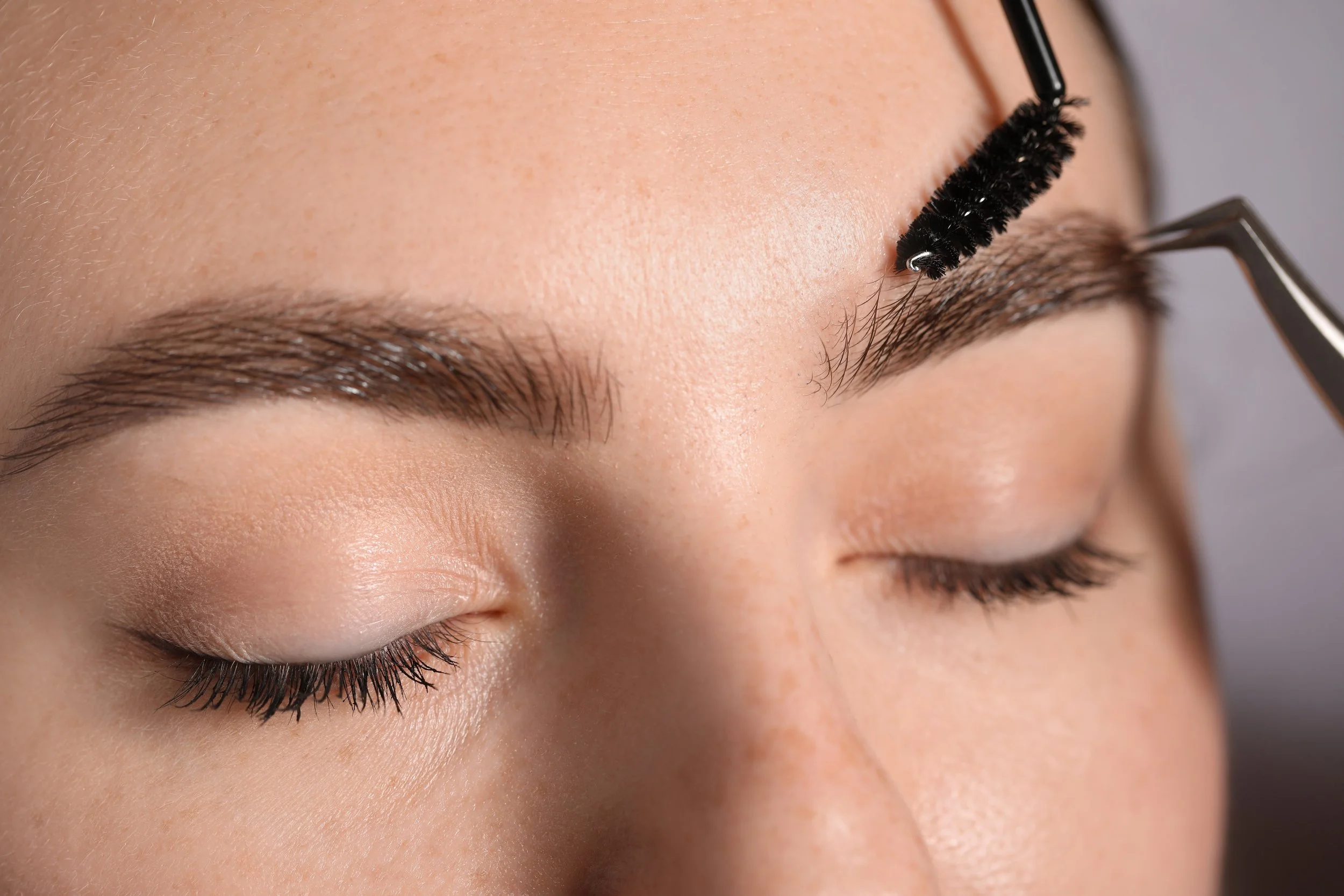 Woman having her eyebrows shaped with a brow brush while lying down with closed eyes and a hand supporting her face.