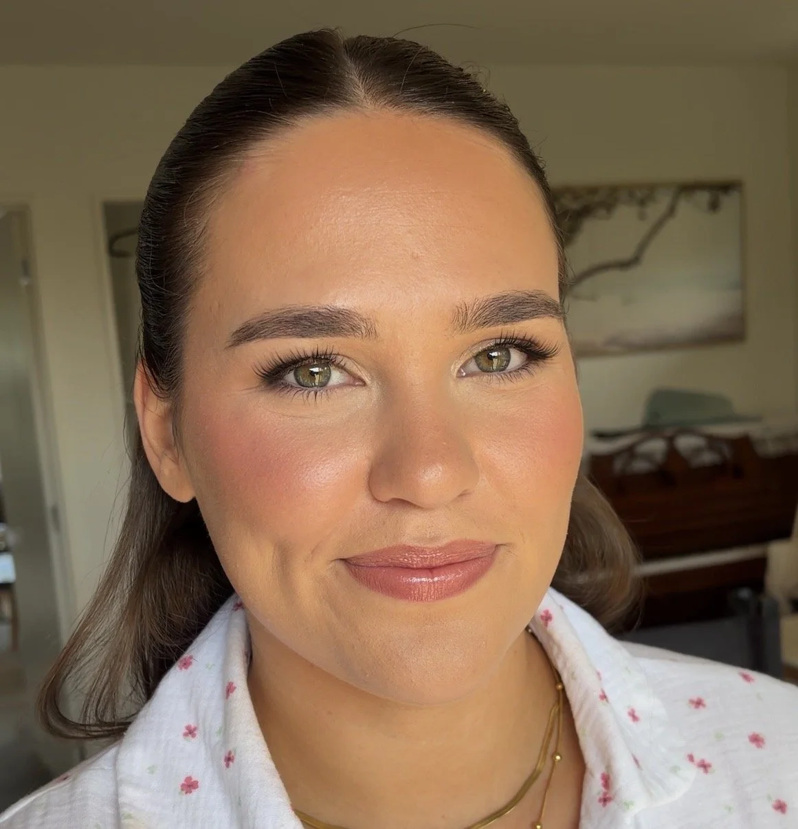 Close-up of a woman with light brown hair, hazel eyes, wearing minimal makeup and a white top with pink floral patterns, in a bright room with a painting on the wall behind her.