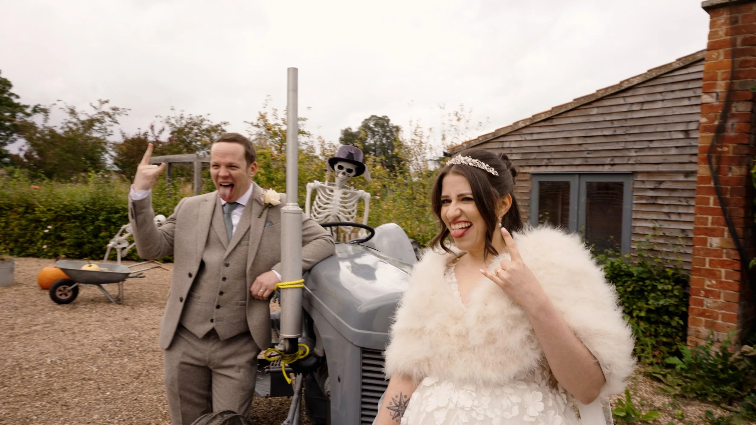 A wedding couple makes silly faces and gestures outdoors with skeleton decorations and a small vehicle in the background.