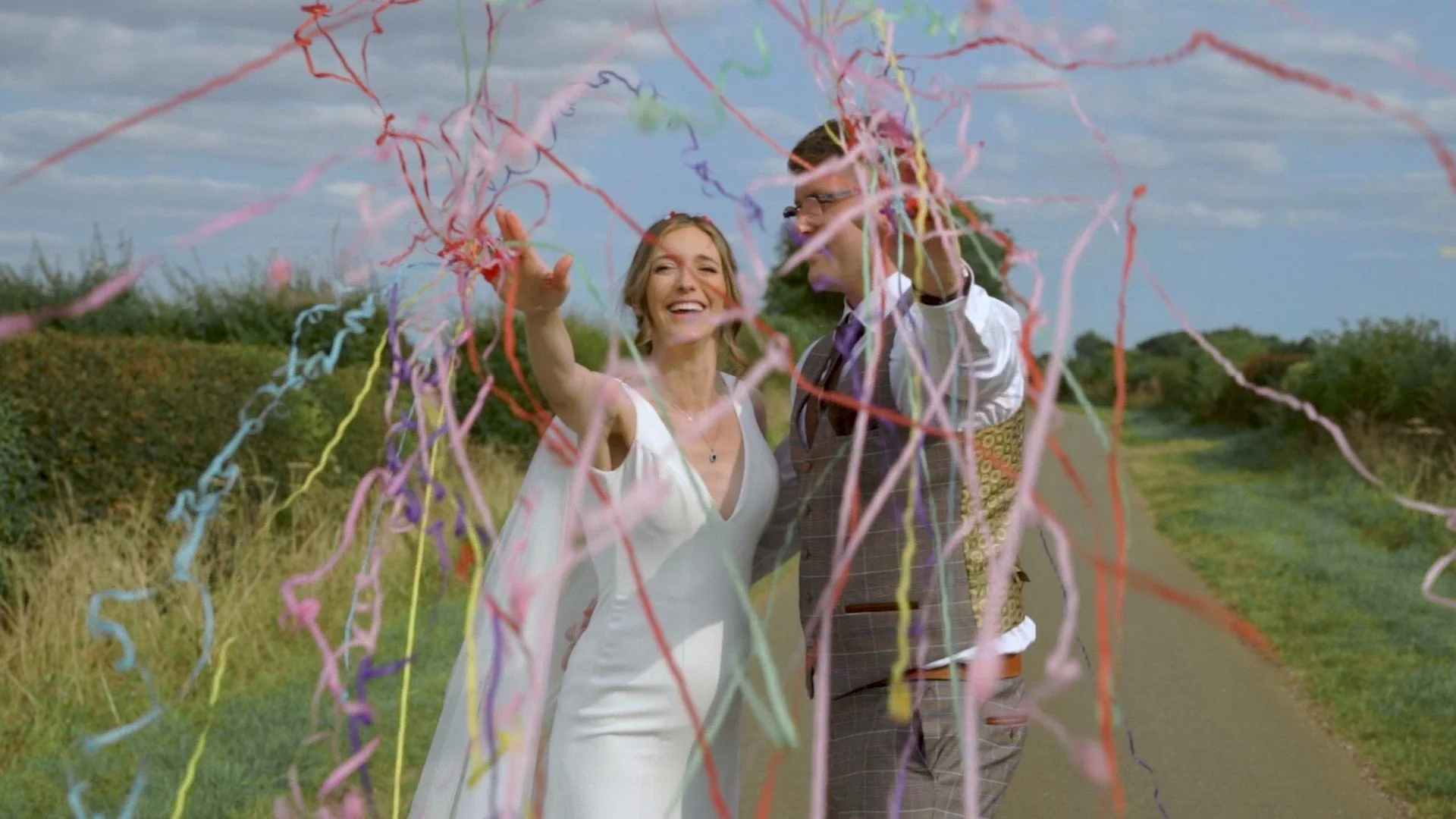 A smiling woman and man celebrating outdoors with colorful streamers in the air, on a country path with grass and bushes on a partly cloudy sky.