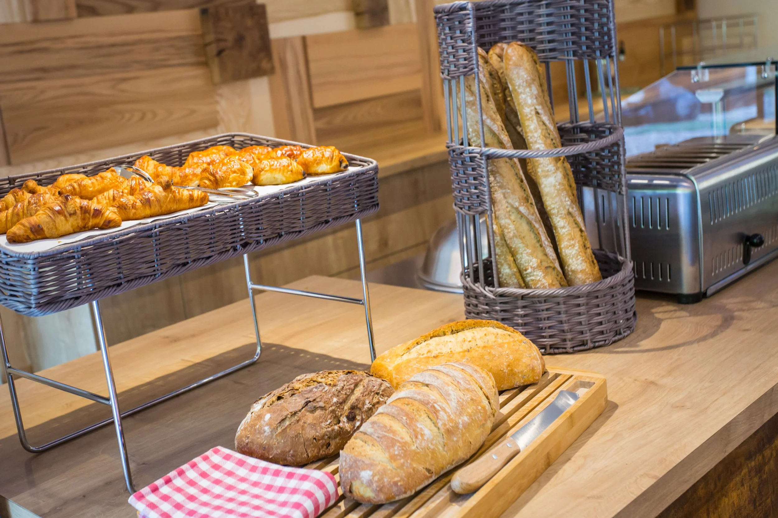 Un assortiment de pains et viennoiseries dans une pâtisserie, incluant des baguettes, un pain complet, et des croissants, avec des toasteurs en arrière-plan.