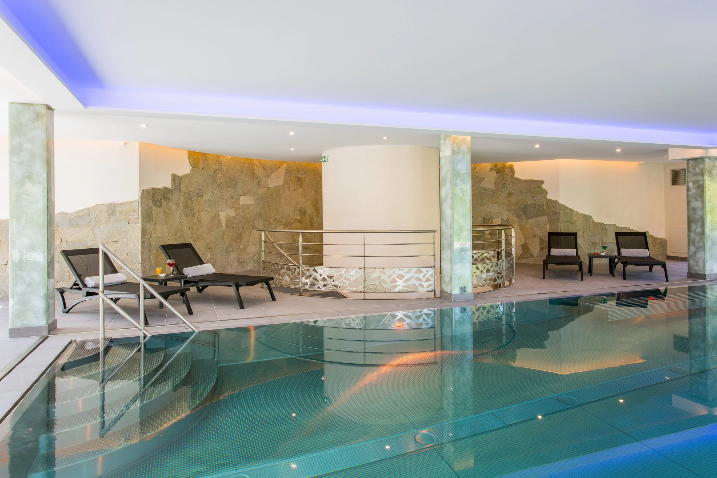 Piscine intérieure dans un spa, avec des chaises longues et des sièges. Mur en pierre et éclairage tamisé.