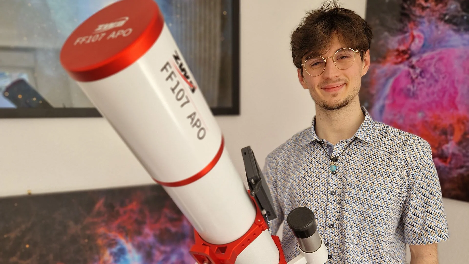 Jeune homme souriant tenant un télescope monté sur un support, portant des lunettes rondes, vêtu d'une chemise à motifs, avec des œuvres d'art astrophotographiques en arrière-plan.